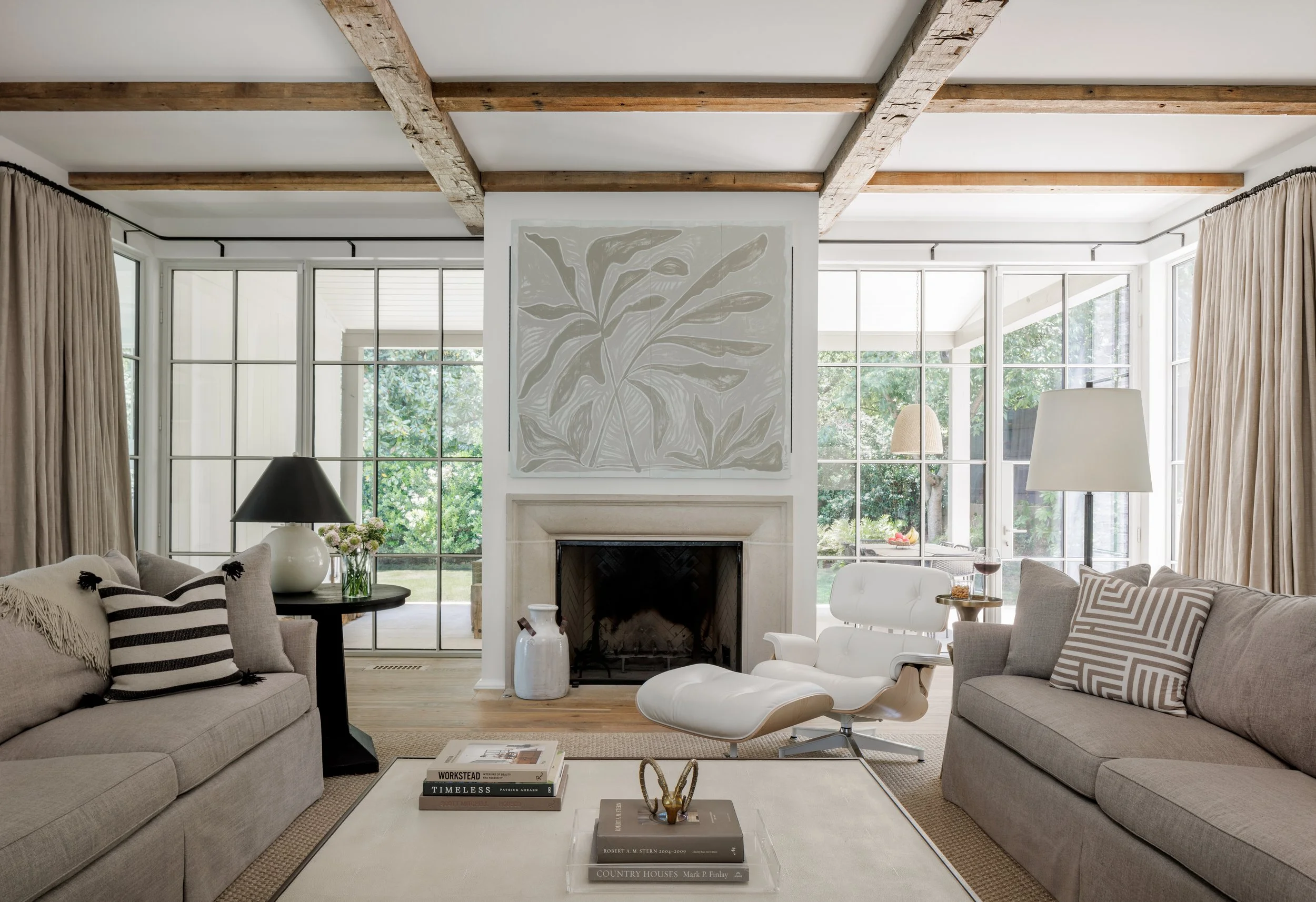 Living room with large windows, beige sofas, a white modern lounge chair, and a fireplace with artwork of leaves above it.