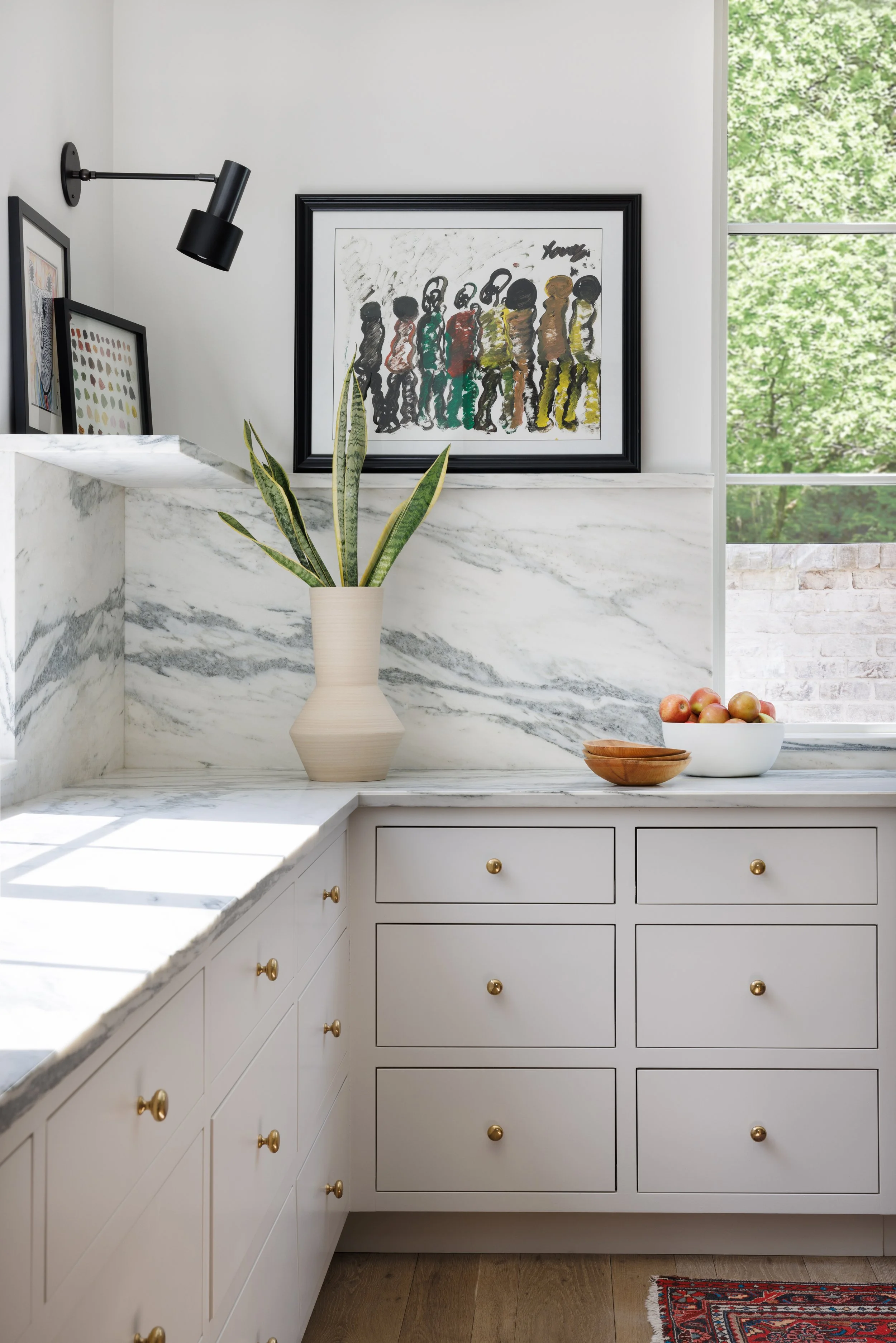 Kitchen counter with white marble surface, green plant in a beige vase, and bowls of apples. White cabinetry with gold knobs. Artwork hanging on the wall and a large window showing green trees outside.