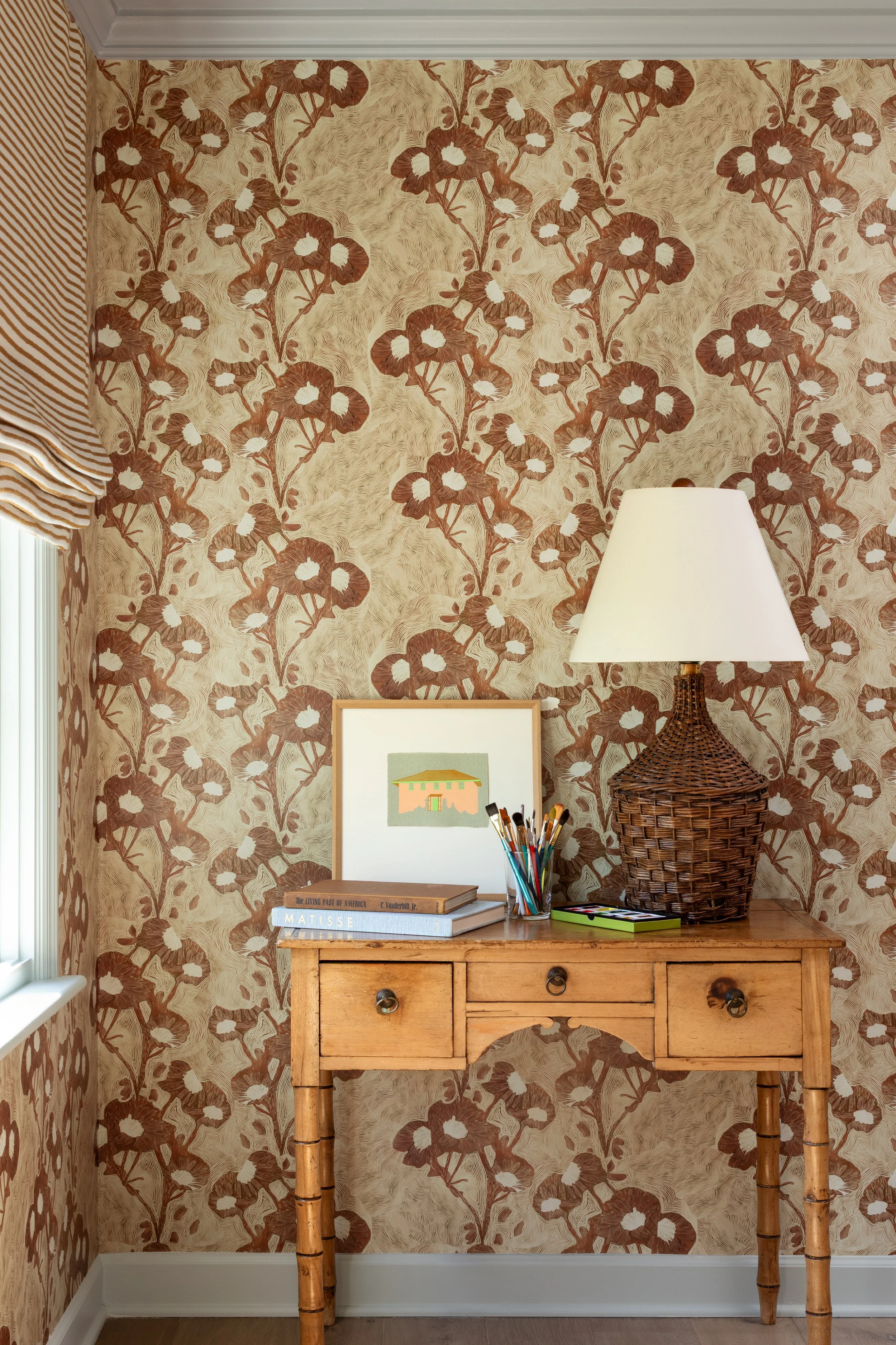 A wooden desk with a woven wicker lamp, a framed picture, books, a cup of pencils, and a small green box, against a floral patterned wallpaper in brown and beige tones.