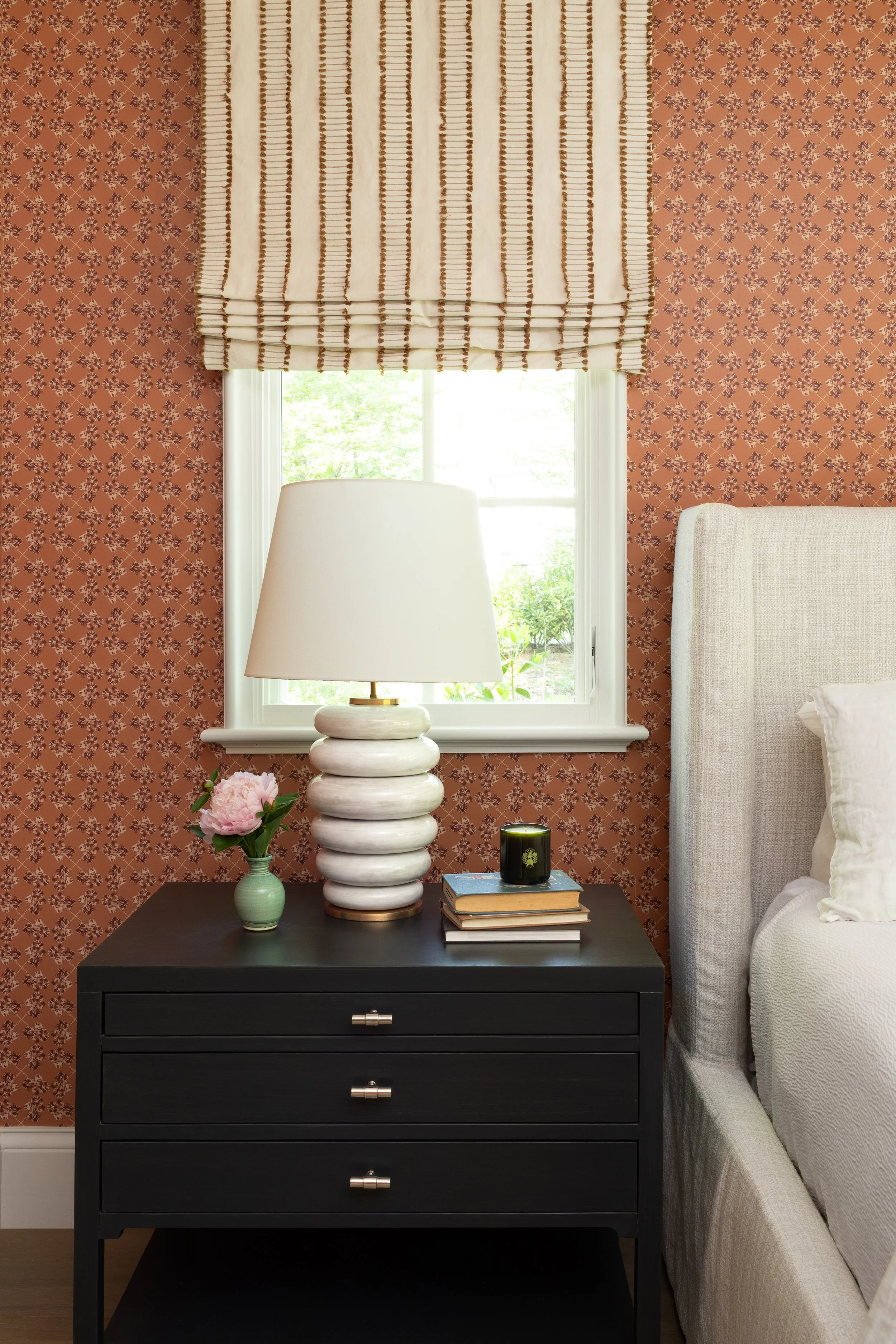 Interior bedroom scene with a black nightstand, a white table lamp, a small green vase with pink flowers, stacked books, a green candle, a window with a beige Roman shade, patterned orange wallpaper, and part of a white upholstered bed and light gray
