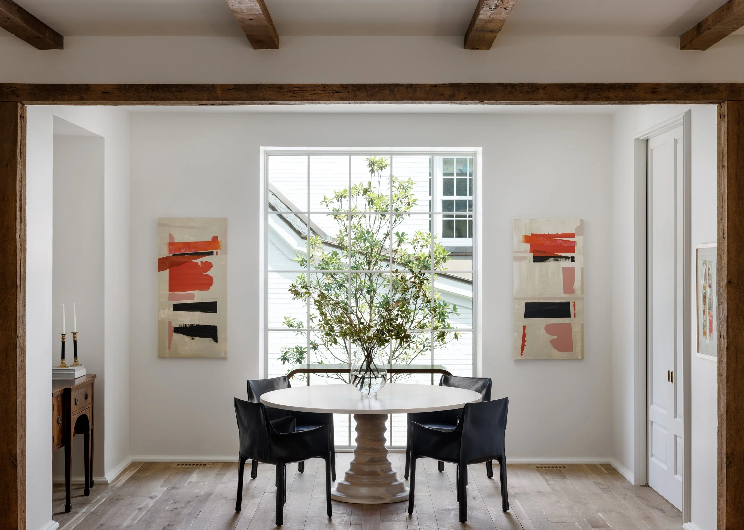 Dining room with a round table, four black chairs, large window with plants outside, artwork on walls, and wooden beams on the ceiling.