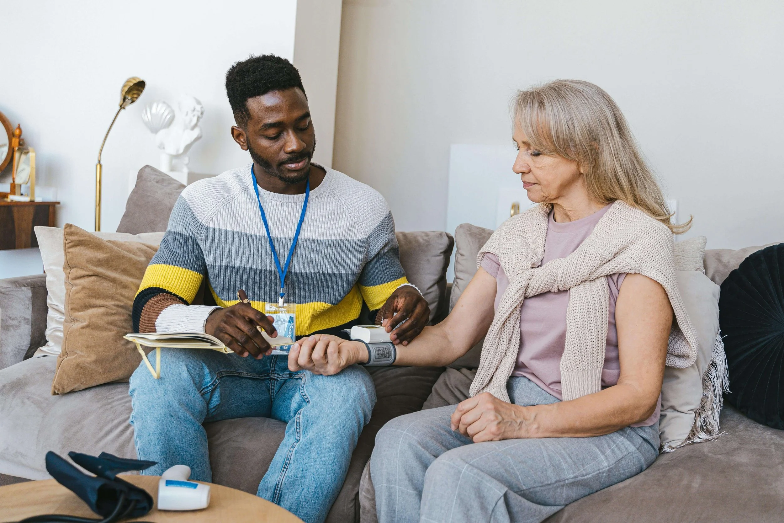 A young man and an older woman sitting on a beige couch, with the man checking the woman's blood pressure using a cuff and stethoscope. The young man is wearing a striped sweater with a lanyard around his neck, and the woman is wearing a light pink shirt and a beige cardigan. They are in a living room with decorative items and a lamp in the background.