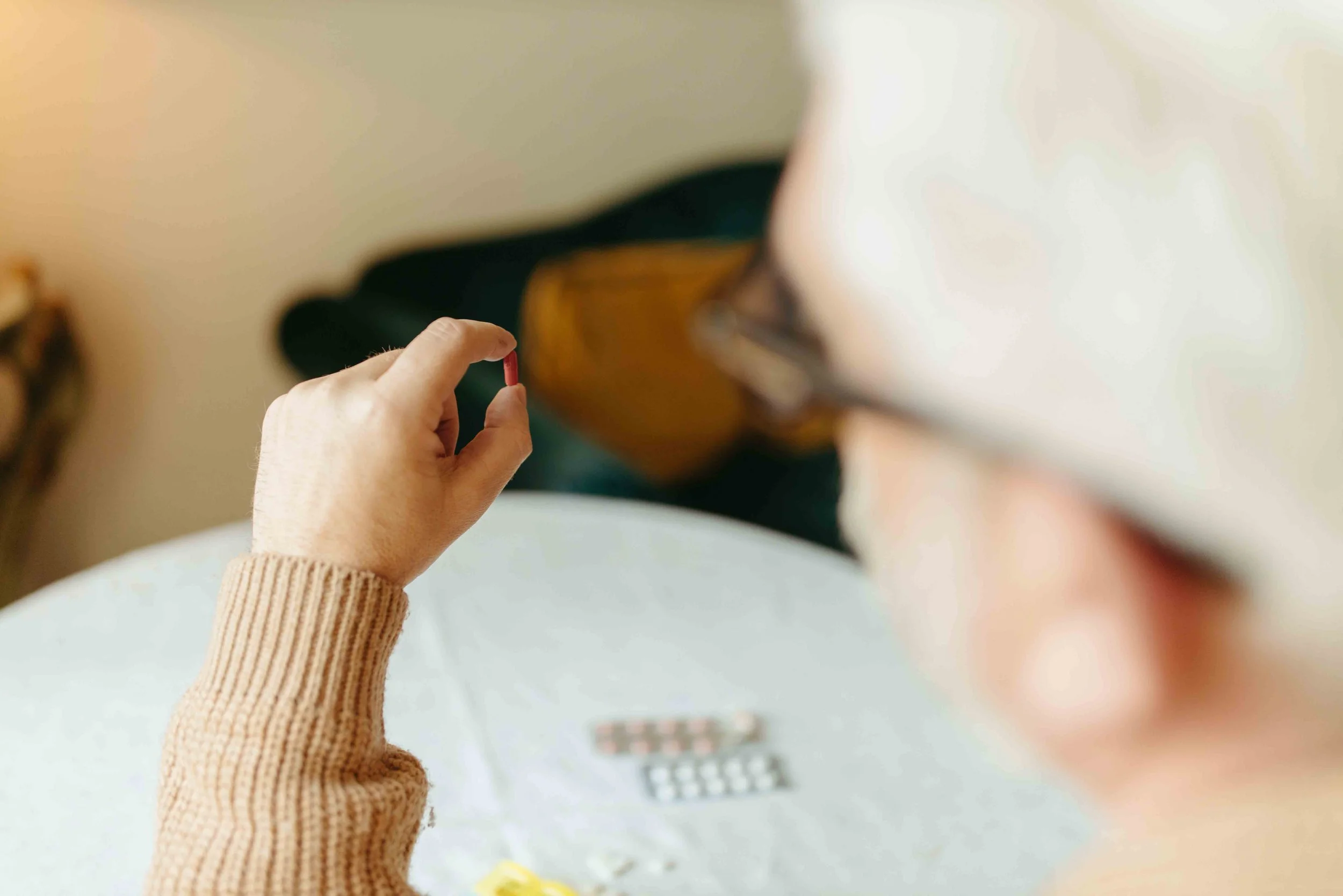 A person wearing a beige sweater holding a red capsule pill between their thumb and index finger.