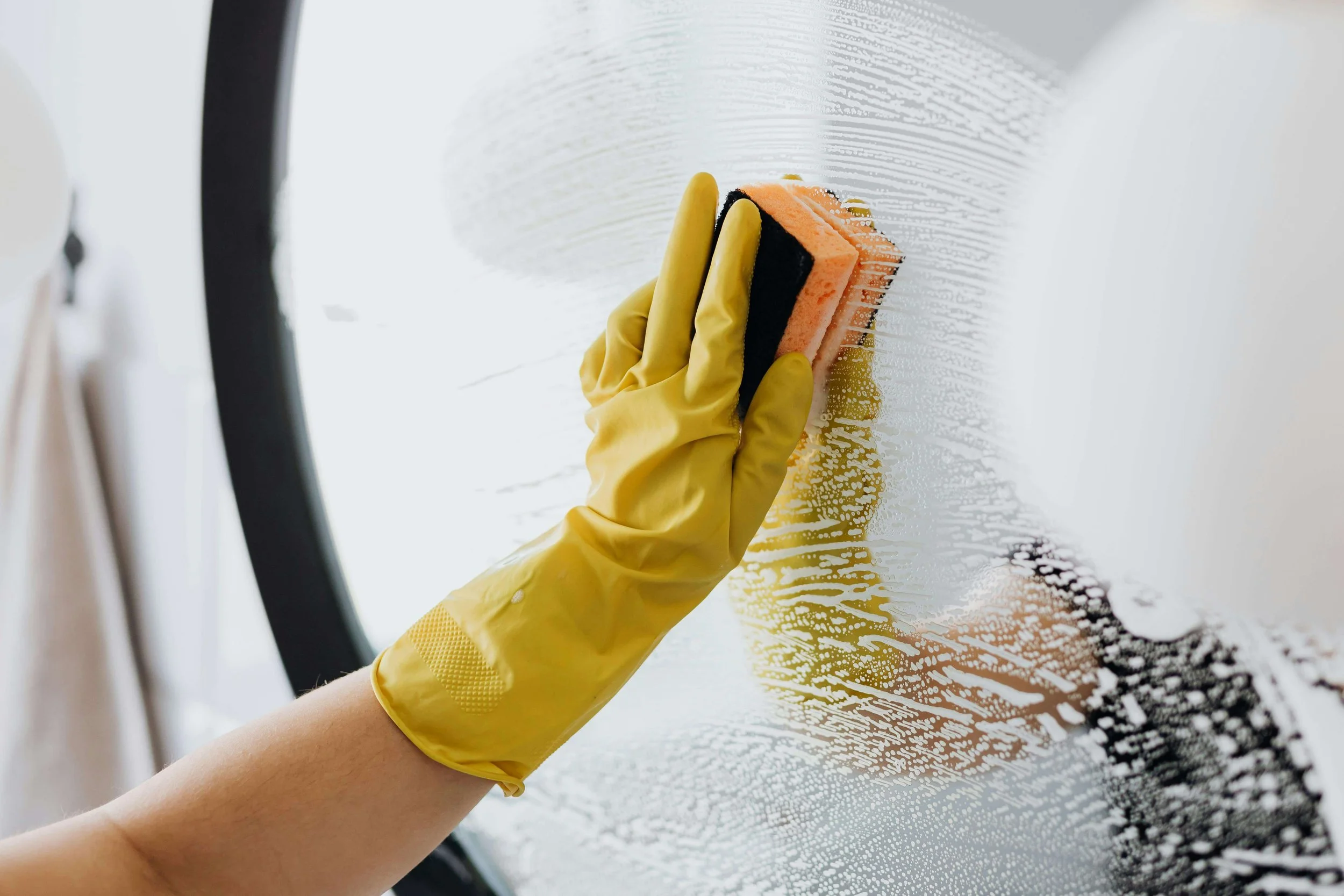 Person wearing yellow gloves cleaning a glass window with a sponge and soap, creating a soapy suds pattern.