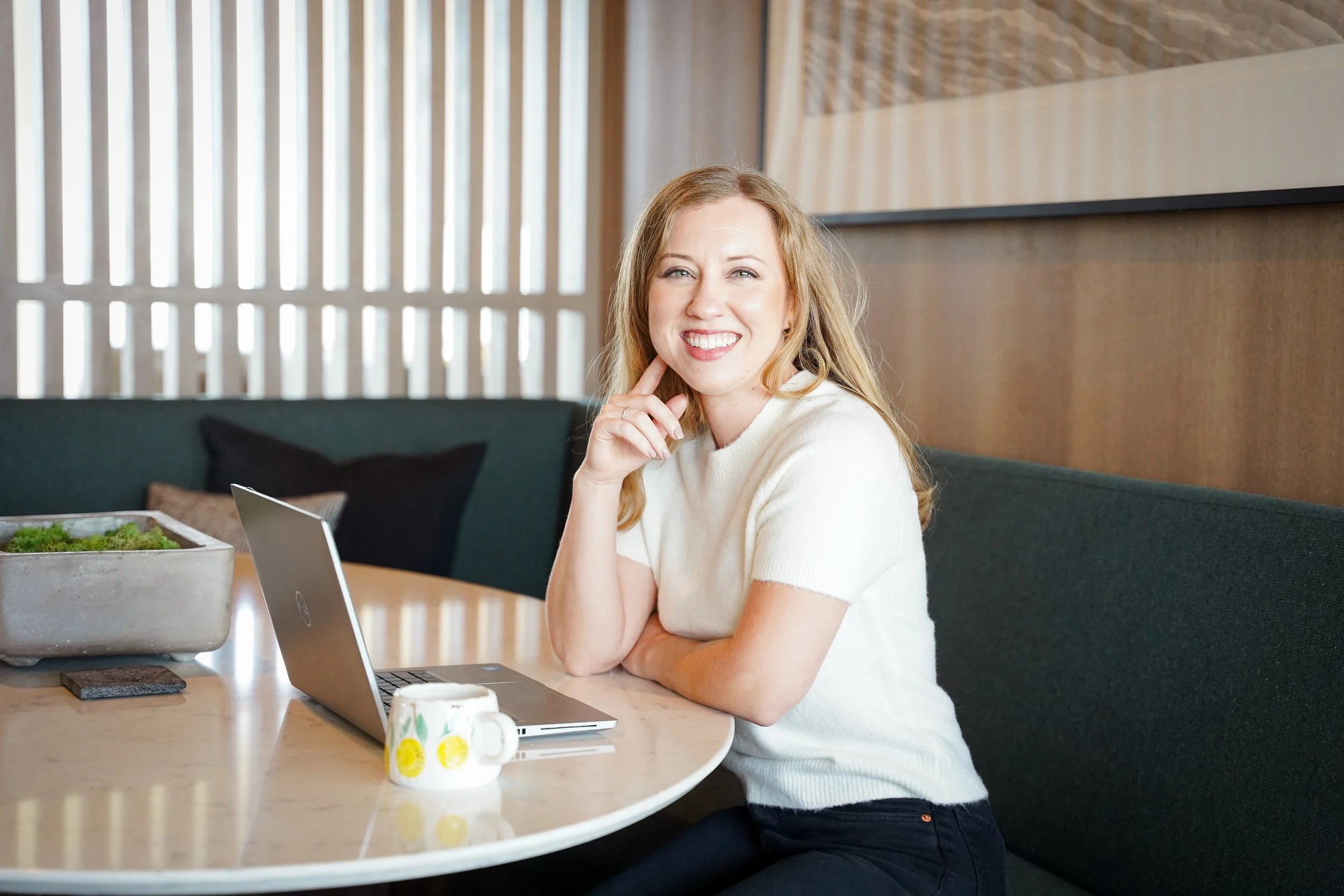 A smiling woman with blonde hair sitting at a table with a laptop and coffee mug.