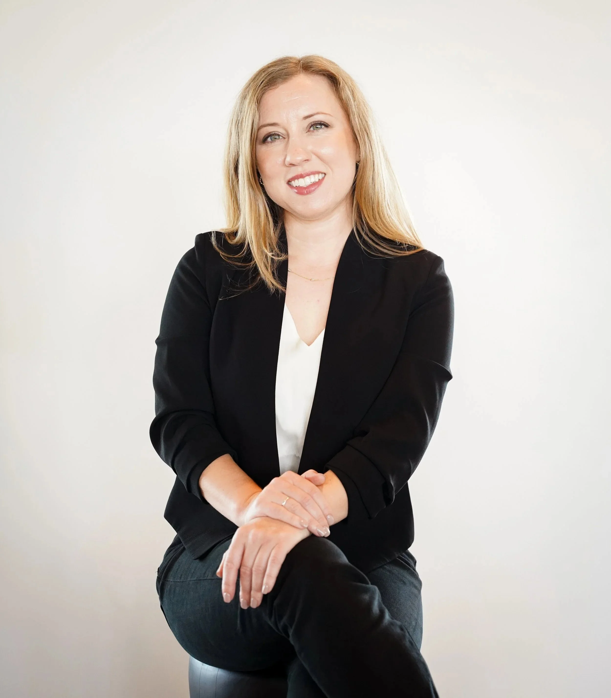 Portrait-style photo of a woman with blonde hair, wearing a black blazer and black pants, sitting against a plain white background.