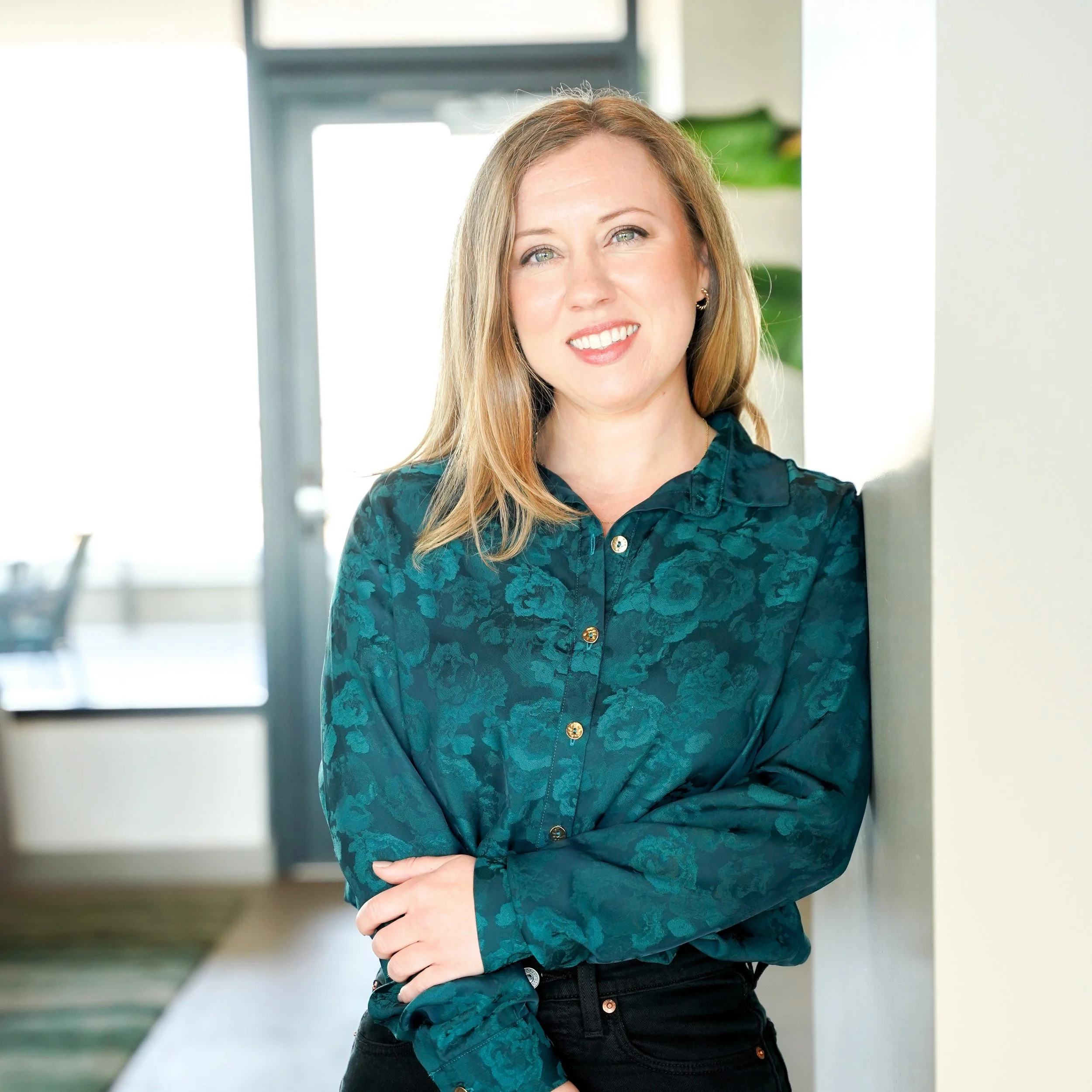 Jessica is a blonde woman wearing a green button-down floral shirt, leaning against a wall and smiling