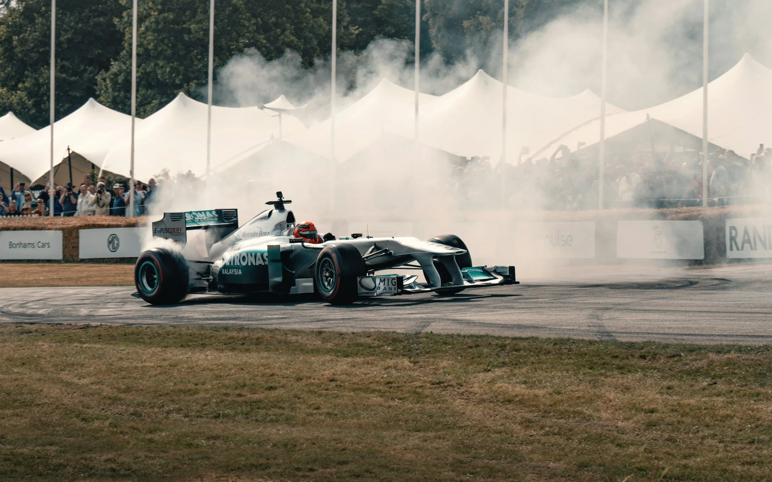 A race car speeds on a track with tire smoke, surrounded by spectators and white tents in the background.