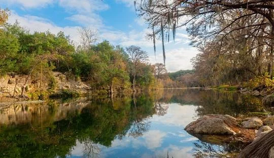 Calm river flowing through a wooded area with rocks along the shoreline and a partly cloudy sky.