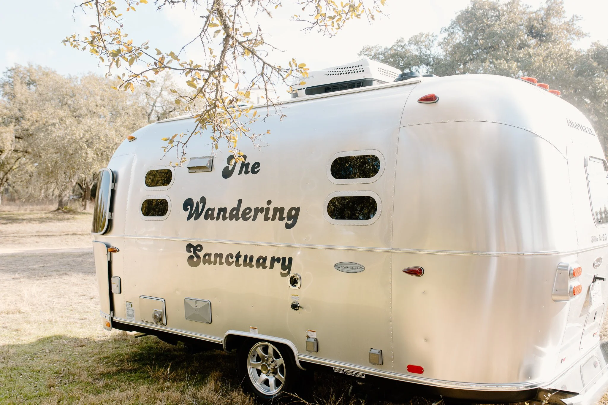 A white camper trailer with black text on the side reading 'the Wandering Sanctuary,' parked on a grassy area with trees and a clear sky in the background.