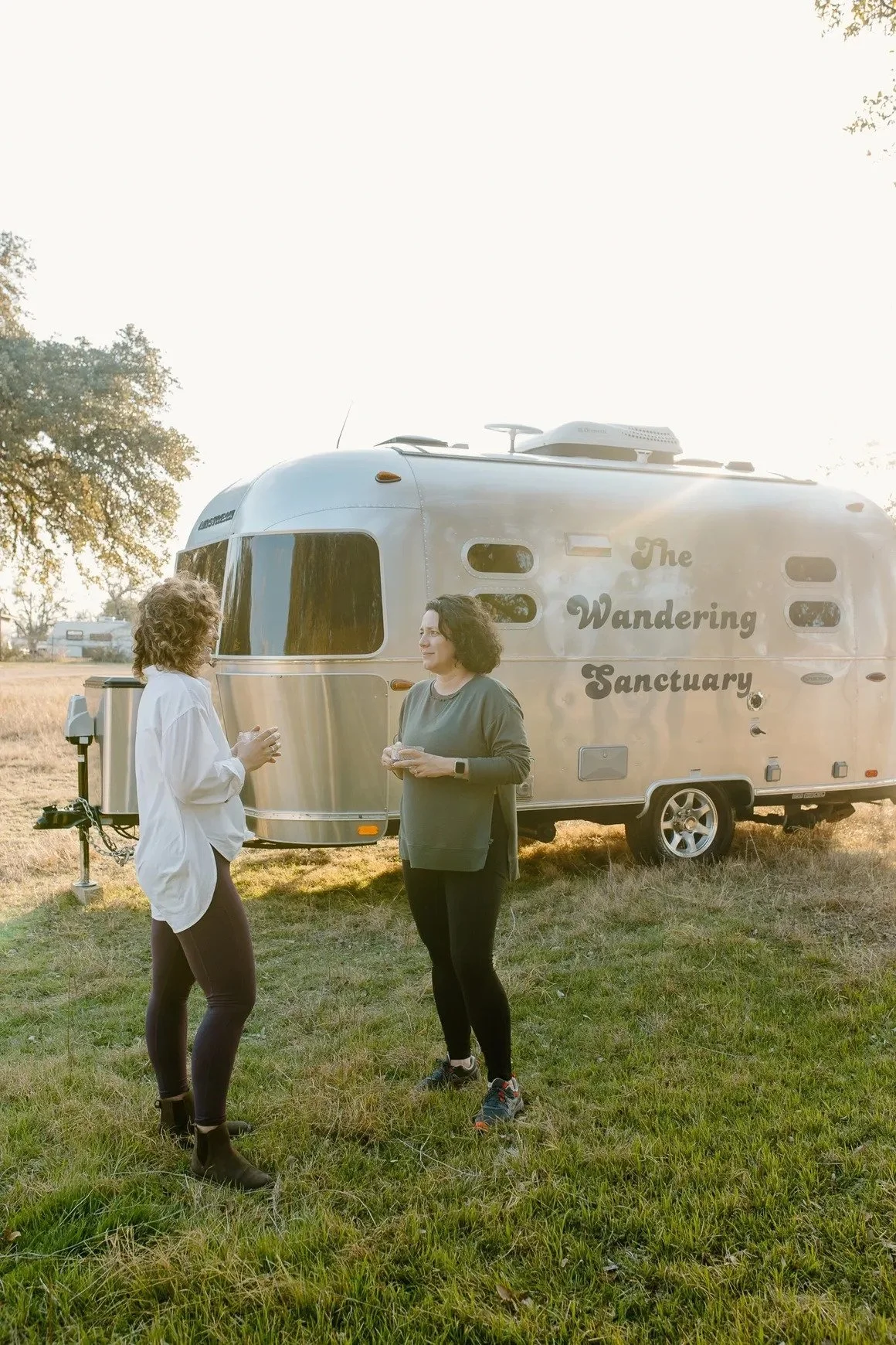 Two women stand and talk outside a silver Airstream trailer labeled 'The Wandering Sanctuary' in a grassy field during sunset, with trees in the background.