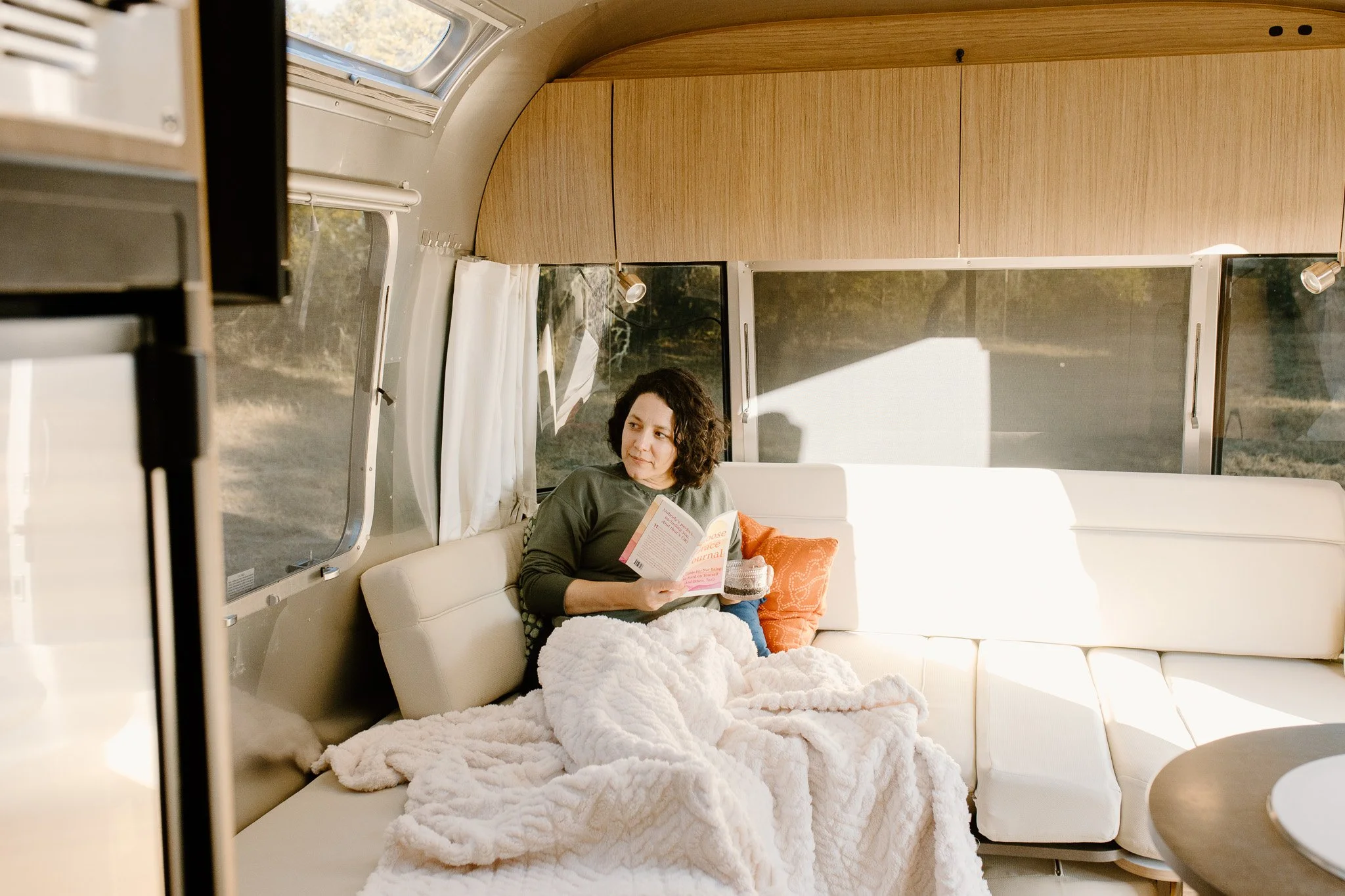 Woman sitting inside a camper van, reading a book, with a blanket on her lap and an orange pillow behind her, during daytime.