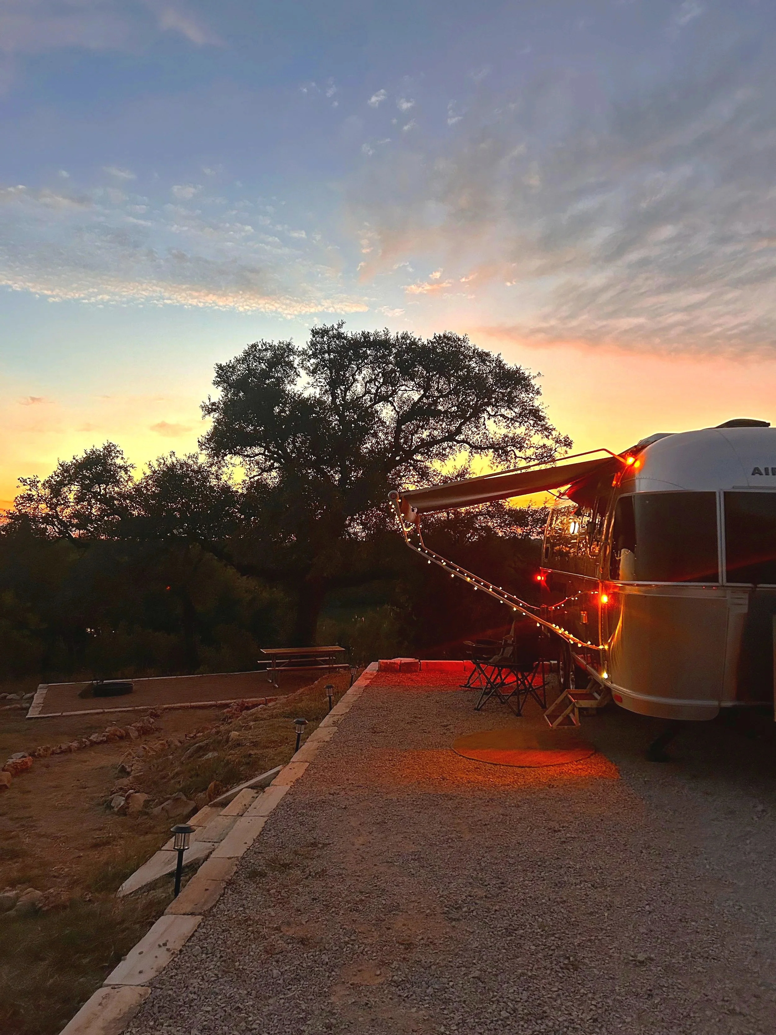 A recreational vehicle parked outdoors at sunset, with string lights hanging from the side, under a sky with clouds and trees in the background.