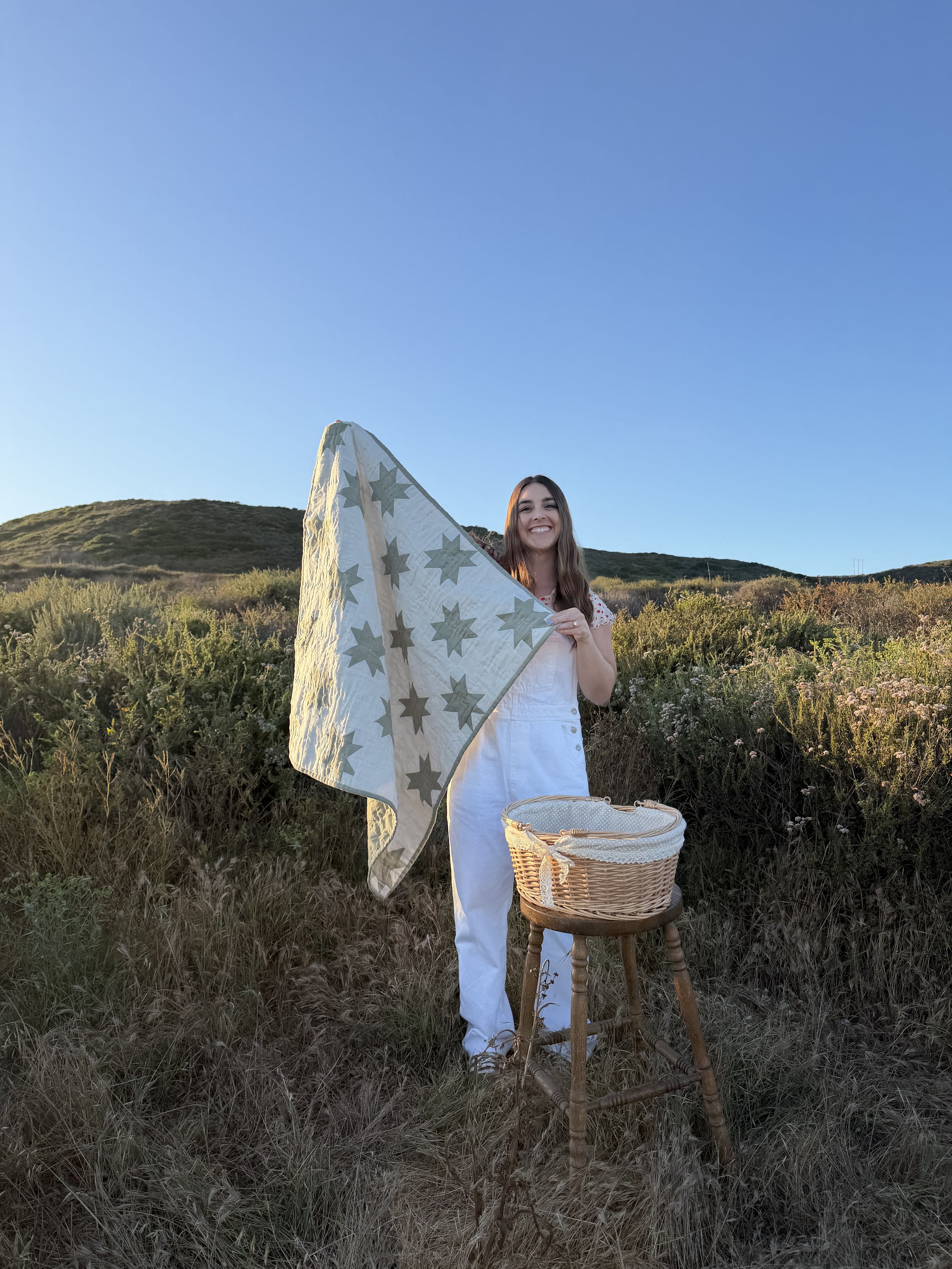 A young woman holding a star-patterned quilt on a stick outdoors in a grassy field at sunset, with a wicker basket on a small wooden stool beside her.