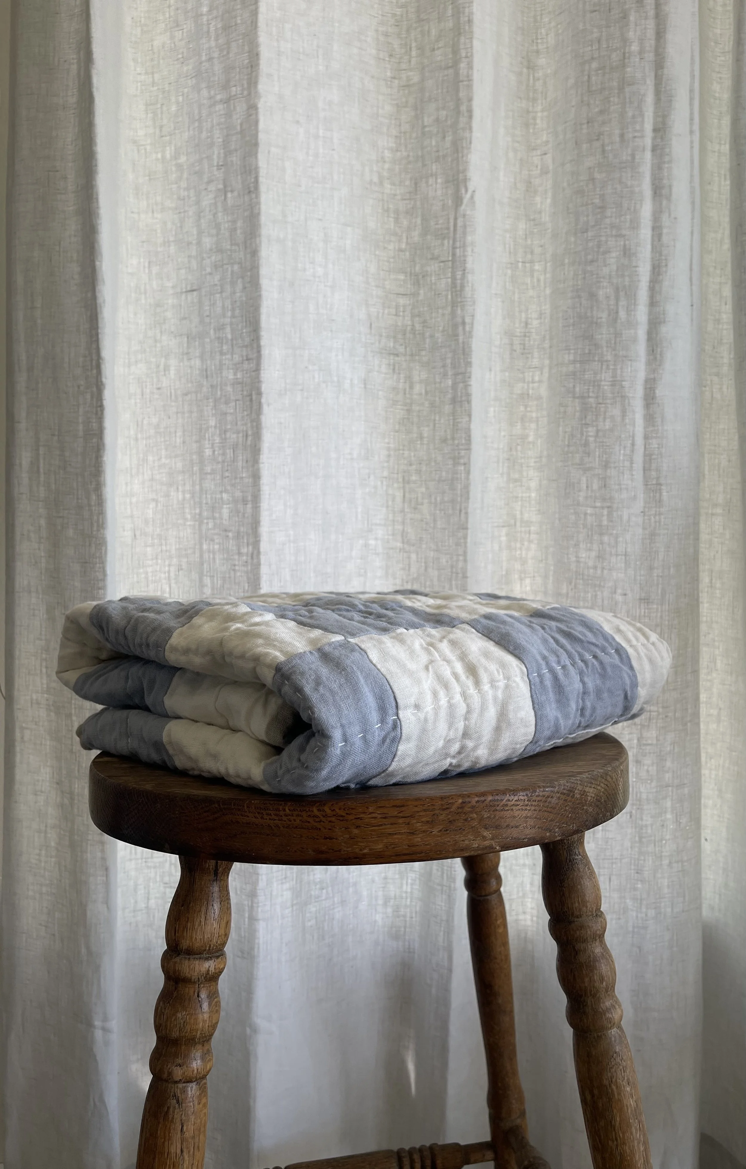 A folded quilt with gray, white, and beige stripes is placed on a wooden stool with turned legs, set against a sheer white curtain background.