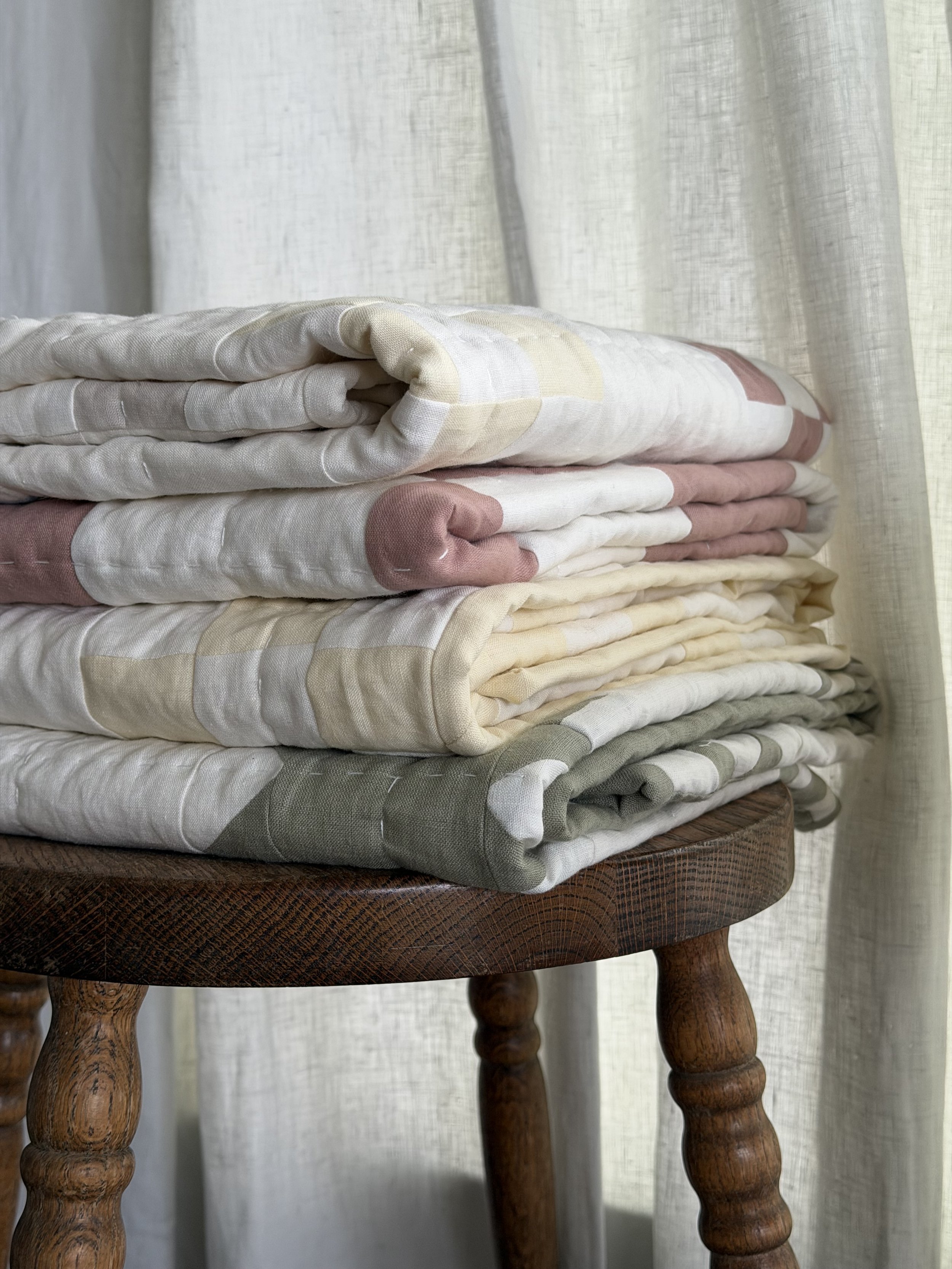 A stack of folded fabric pieces in neutral colors, placed on a round wooden table with carved legs, behind a light-colored curtain.