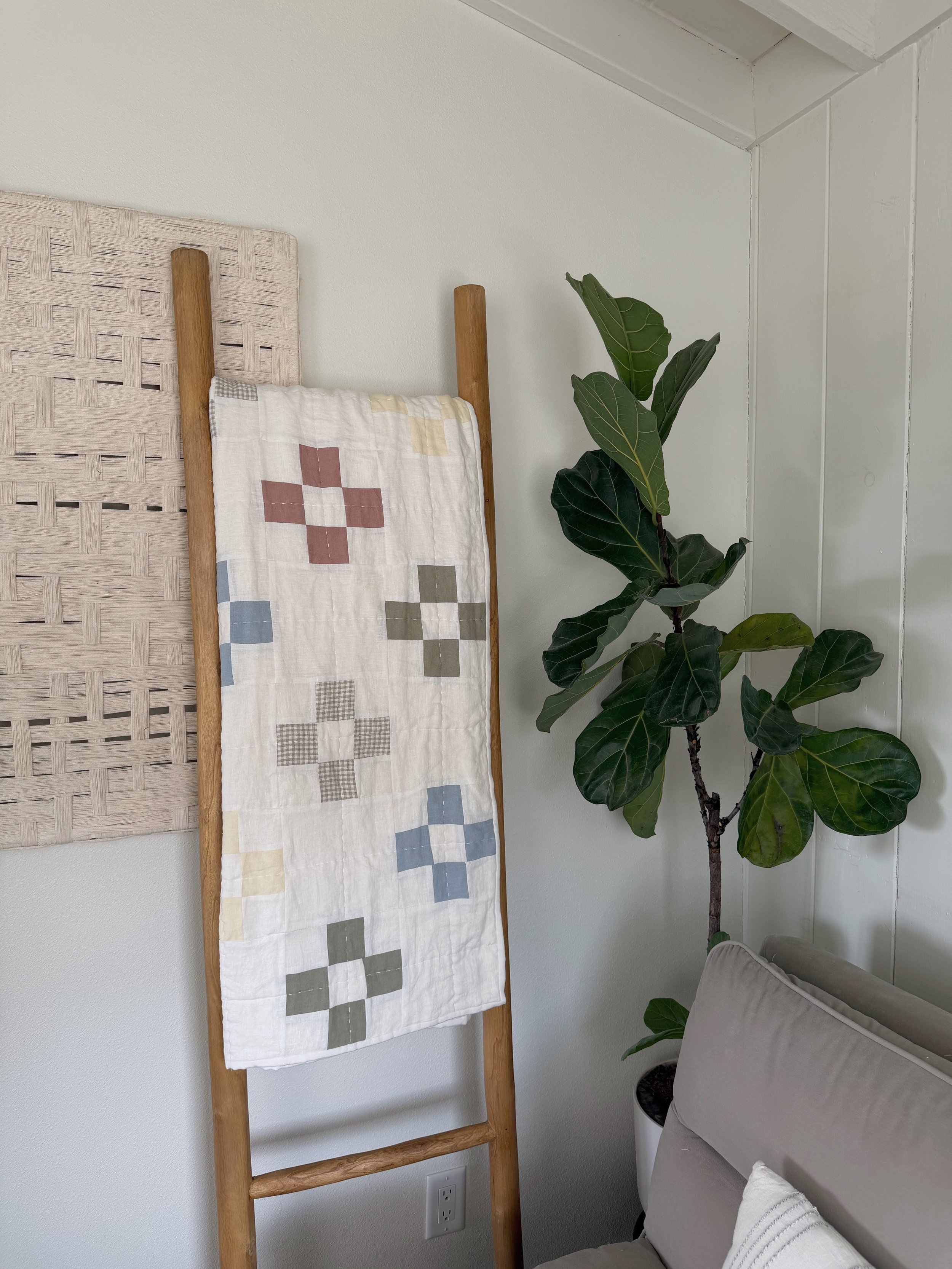 A corner of a room with a wooden ladder-style rack, a quilt with a patchwork pattern hanging on it, a large green potted fiddle leaf fig plant, part of a beige armchair, and a white wall with a textured panel.