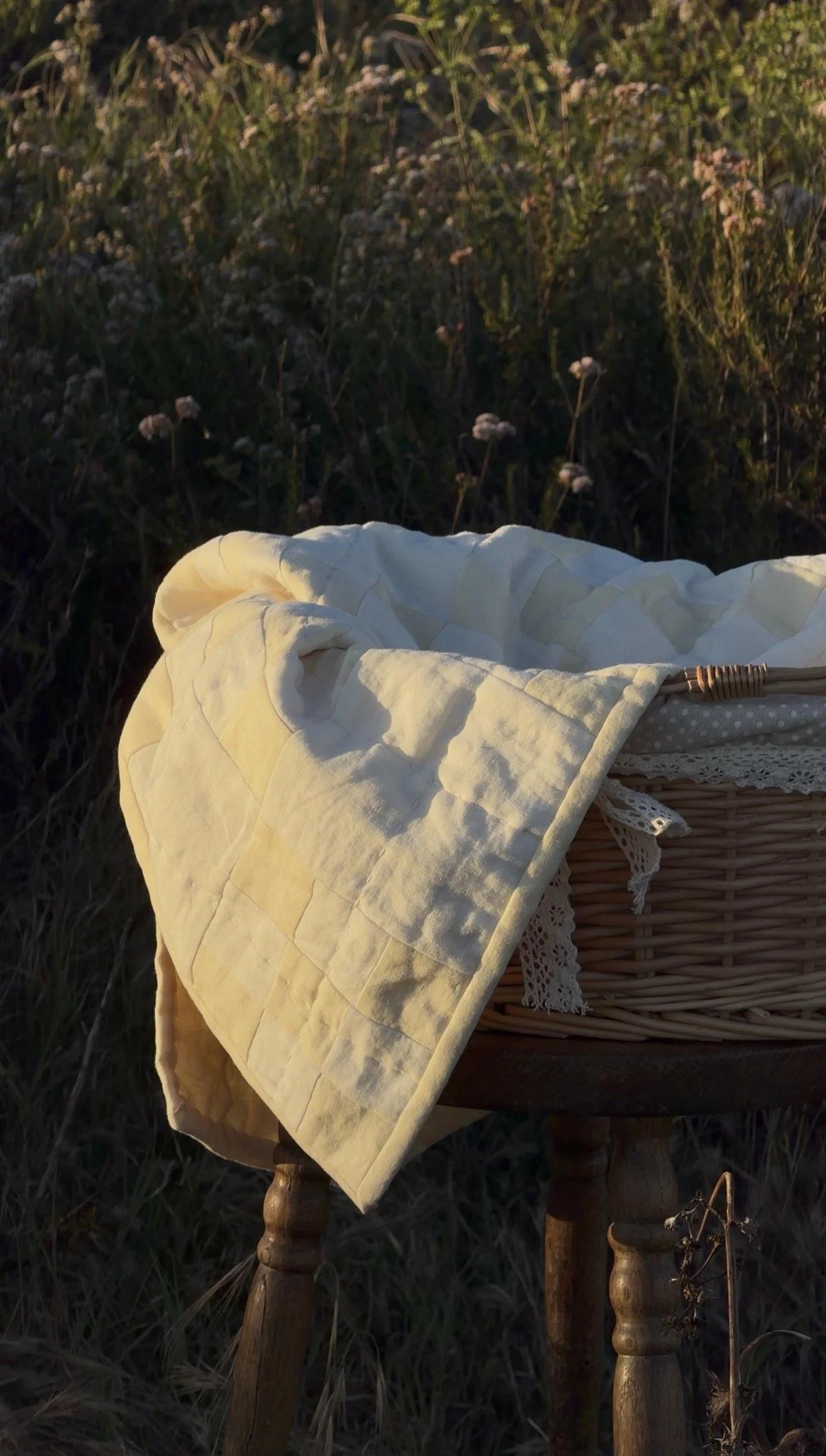 A woven basket with a cream-colored quilt draped over it, set outside on a wooden table in a grassy field during late afternoon or early evening.