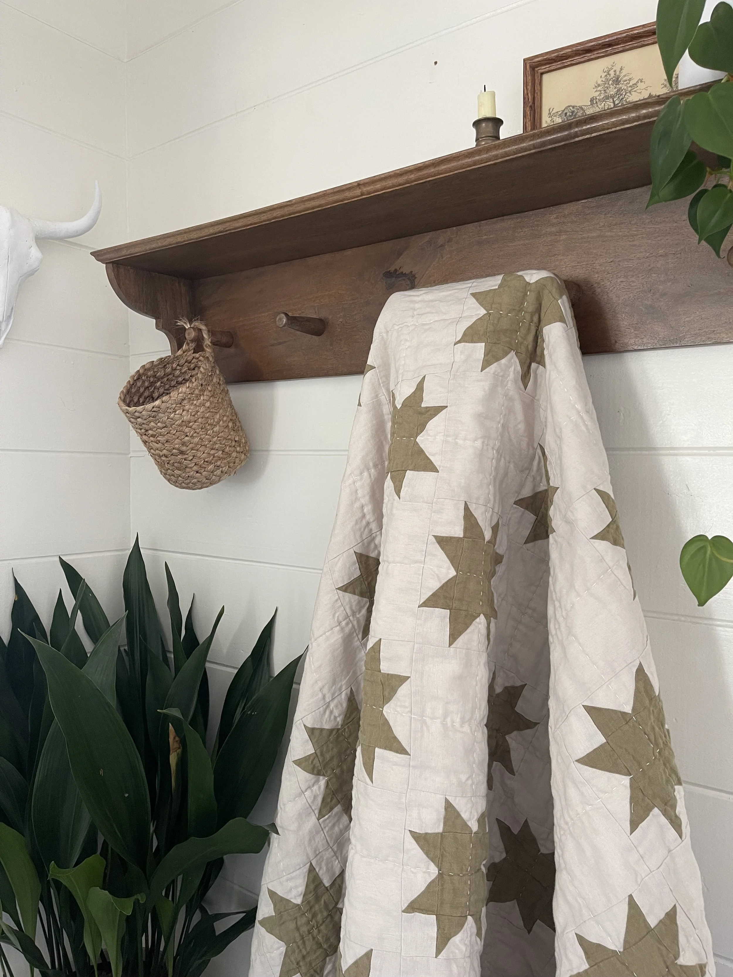 A decorative wall hanging with a wooden shelf, a woven basket, a quilt with leaf pattern, and plants, mounted on a white wall.