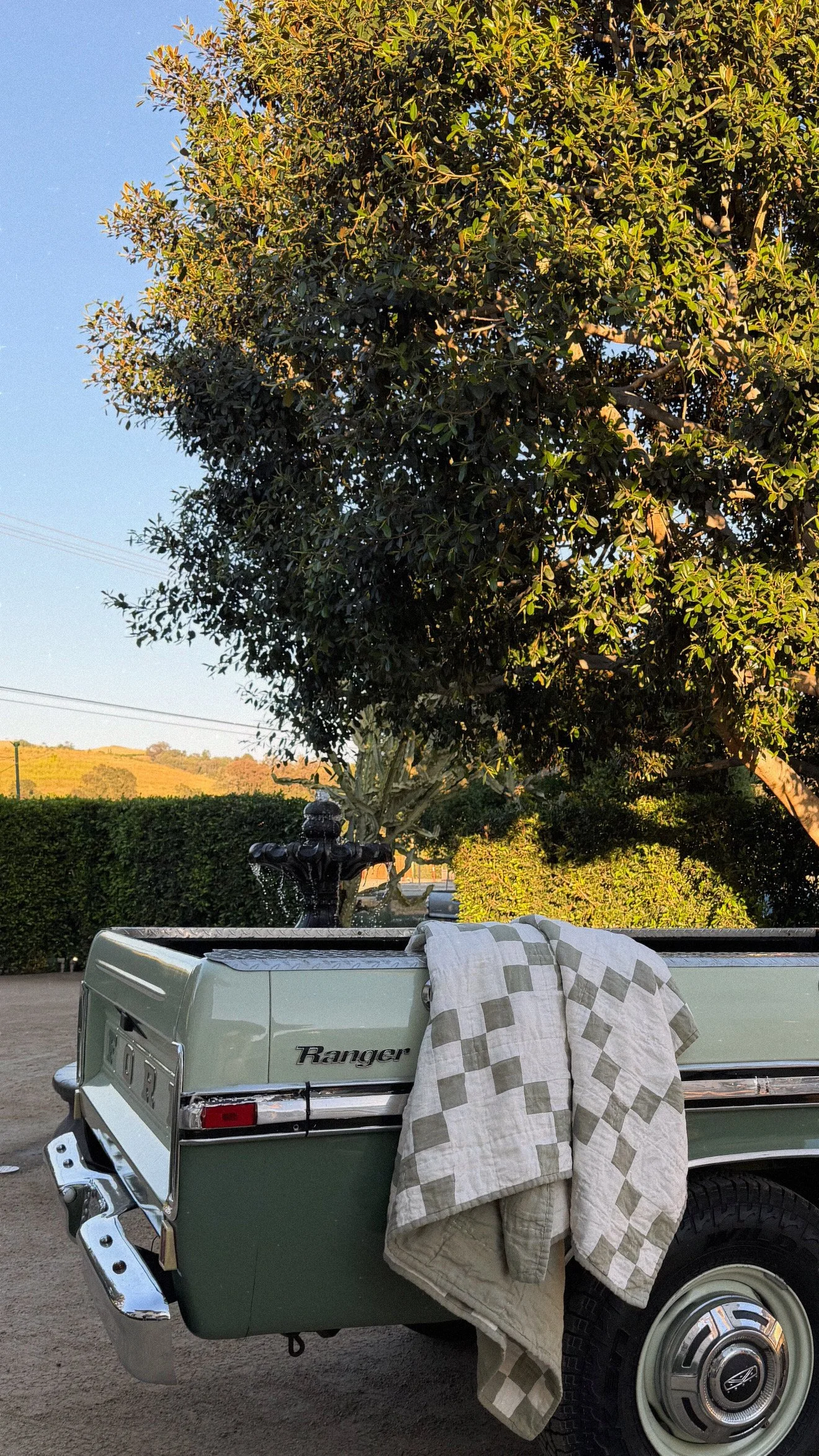 Close-up of a vintage green Ford Ranger pickup truck with a beige plaid blanket draped over its tailgate, parked outdoors near a hedge, trees, and a fountain, with hills in the background during daylight.