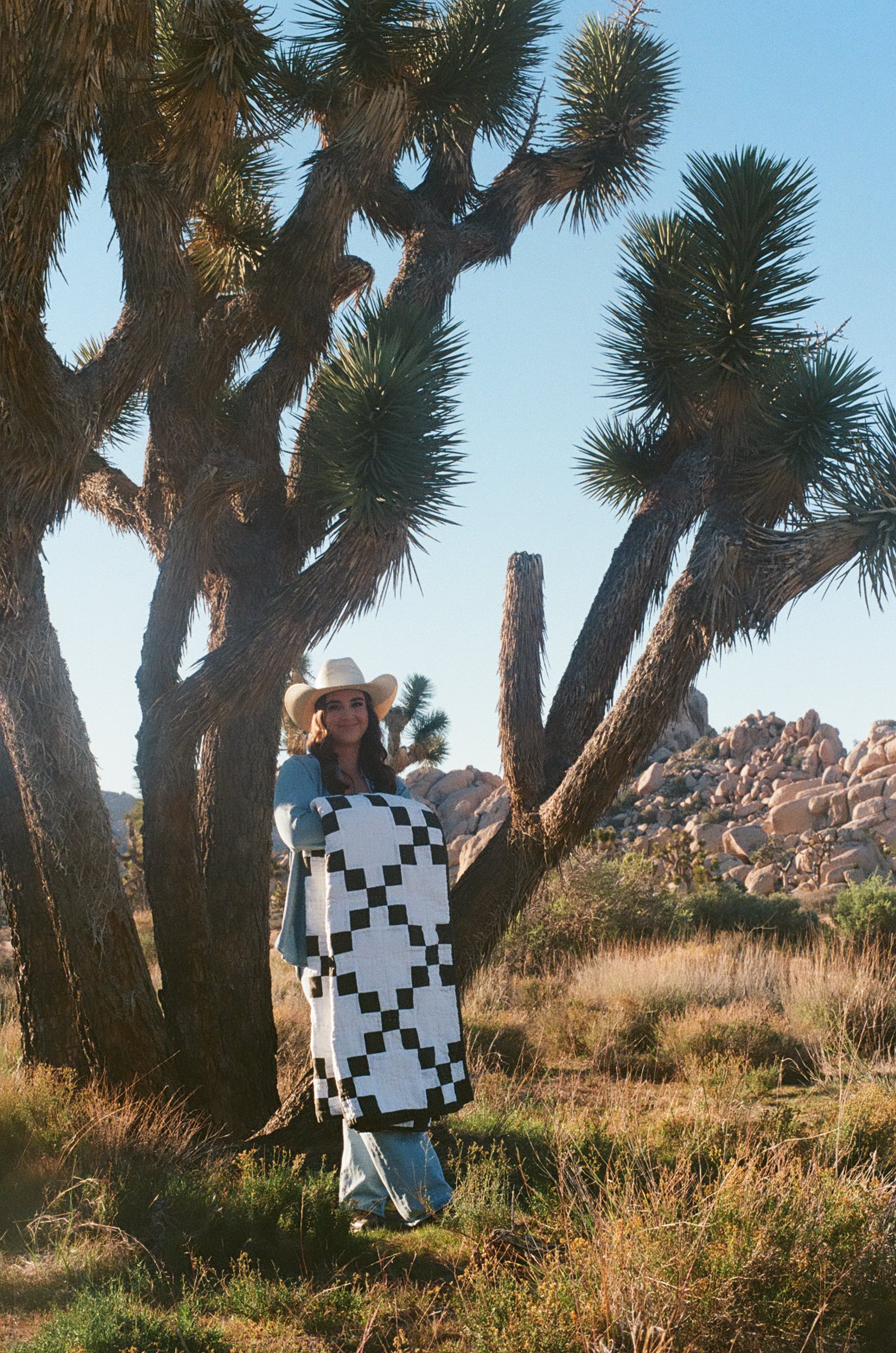 A woman wearing a wide-brimmed hat and a black-and-white checkered blanket dress stands outdoors among desert plants, with Joshua trees and rocky terrain in the background.