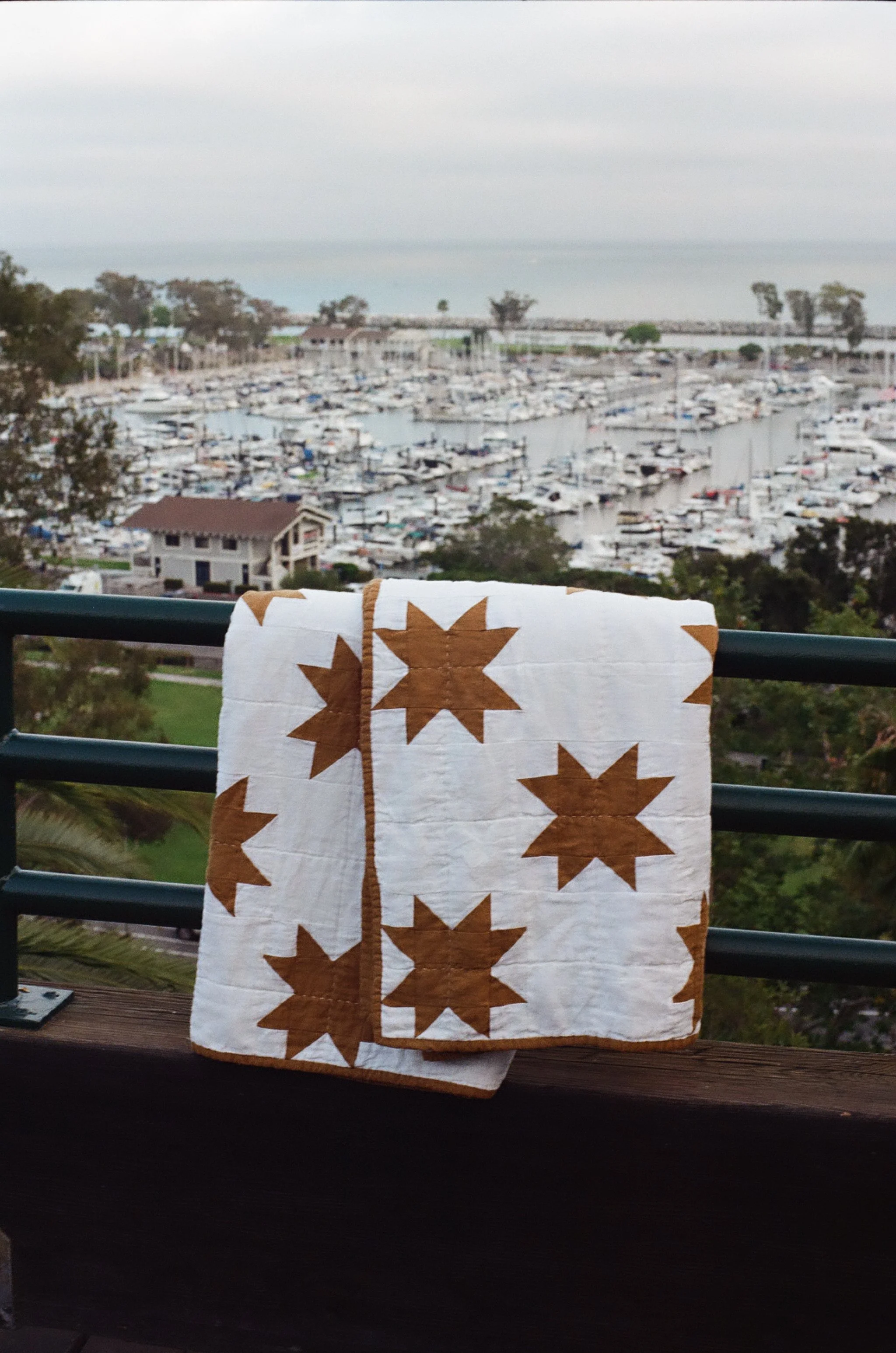 A landscape view of a marina with sailboats and yachts docked in water, with trees and a bridge in the background, seen from a balcony with a quilted fabric hanging on a railing.