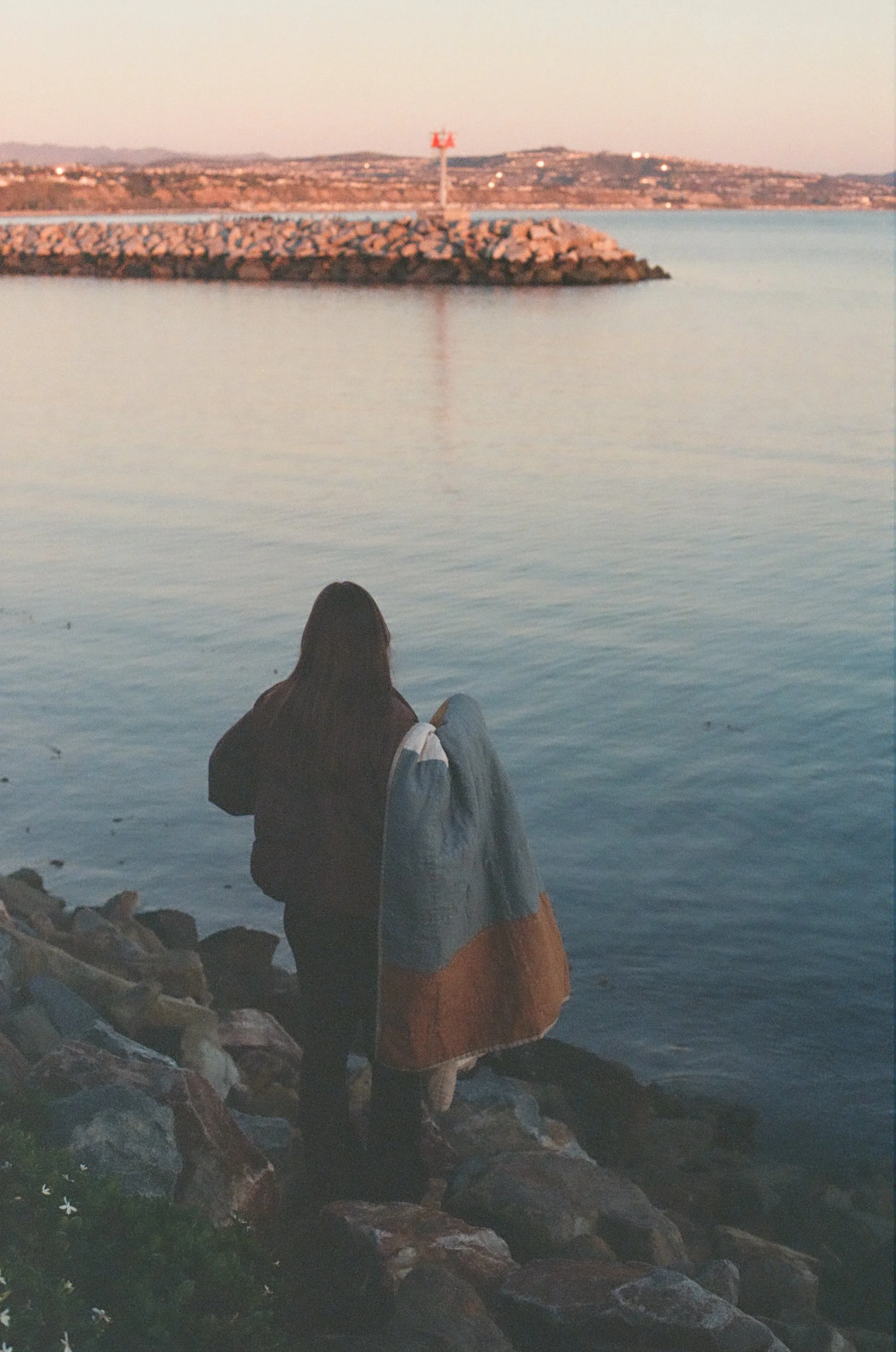 Person sitting on rocks by the water, facing the shore with a breakwater and distant land in the background during sunset.