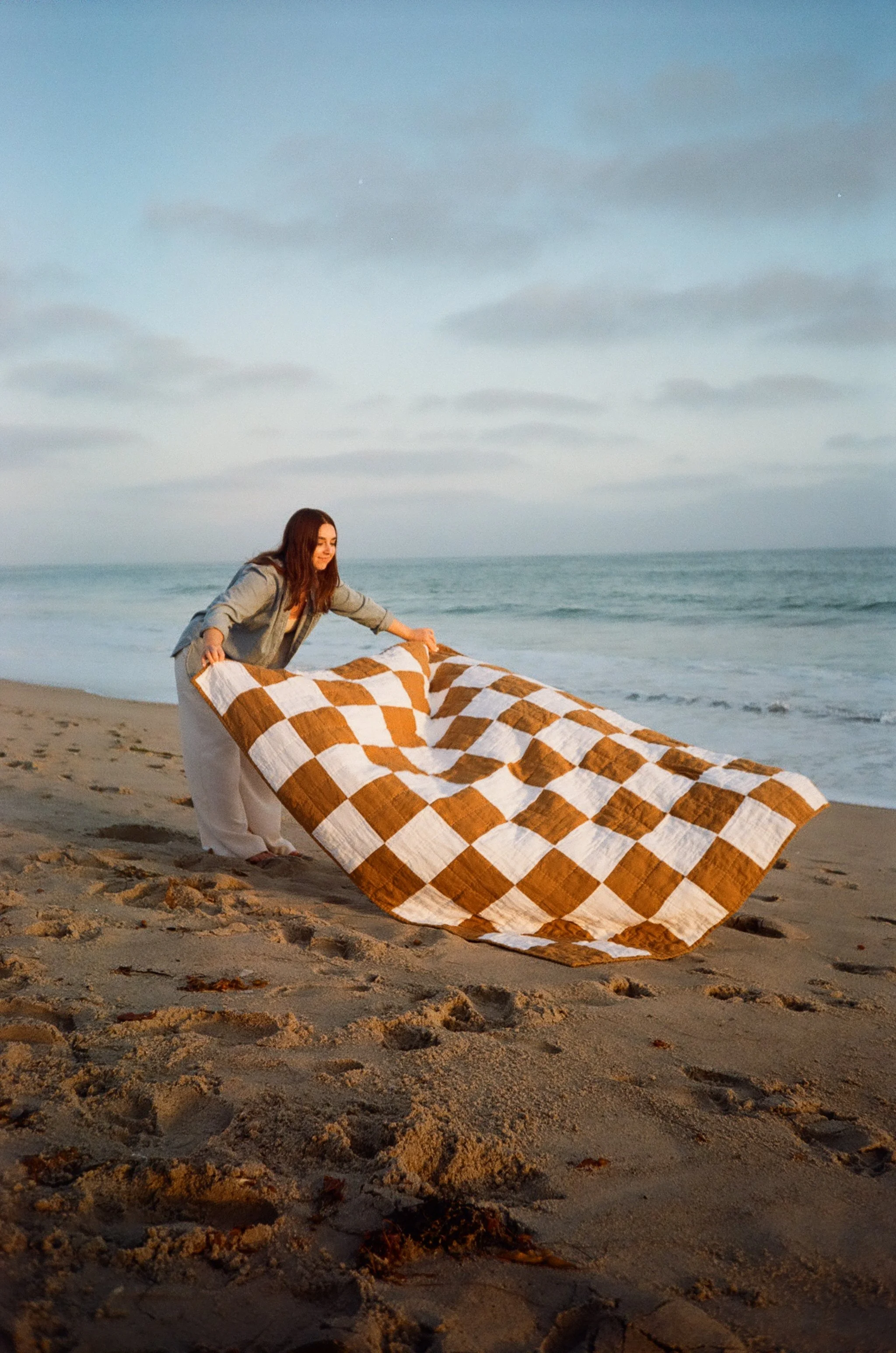 A woman on a beach holding a large checkered blanket, with the ocean and cloudy sky in the background during sunset or sunrise.