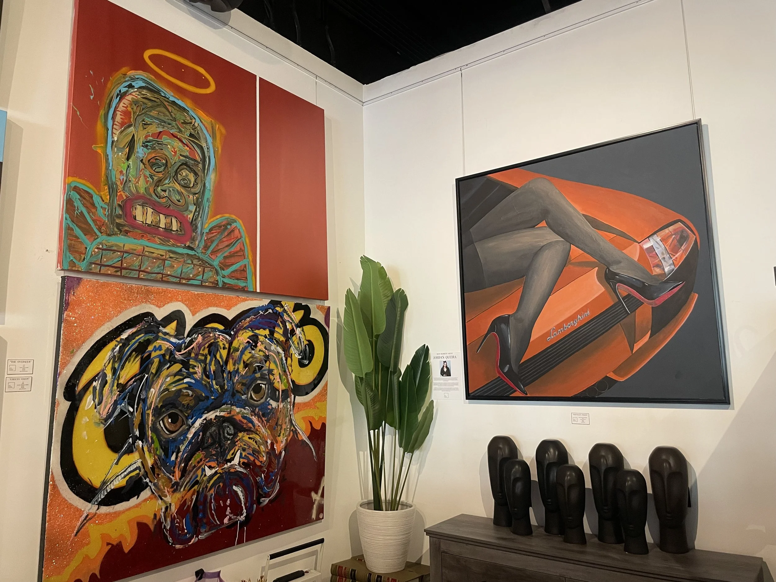 Art gallery displaying colorful abstract and portrait paintings, with a potted plant and black chairs in the scene.