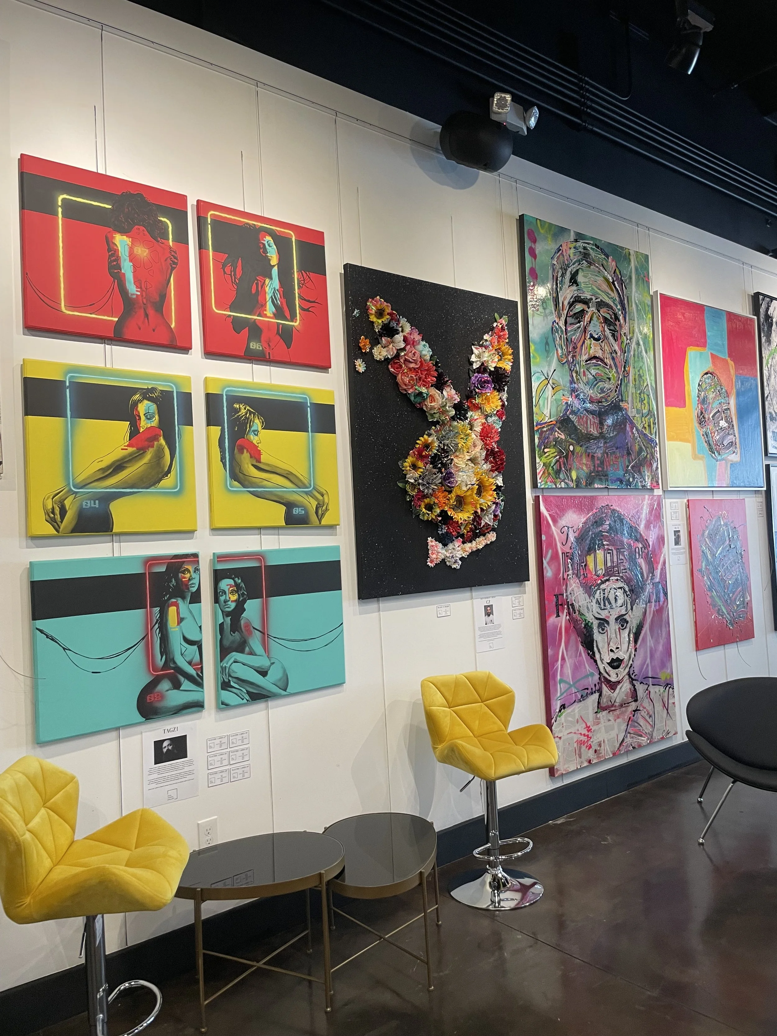 Art gallery wall displaying several colorful paintings, including pop art portraits with neon outlines, a black textured piece with 3D floral 'V' shape, and a large portrait of a woman with vibrantly painted features. Yellow velvet chairs and small tables are in the foreground.