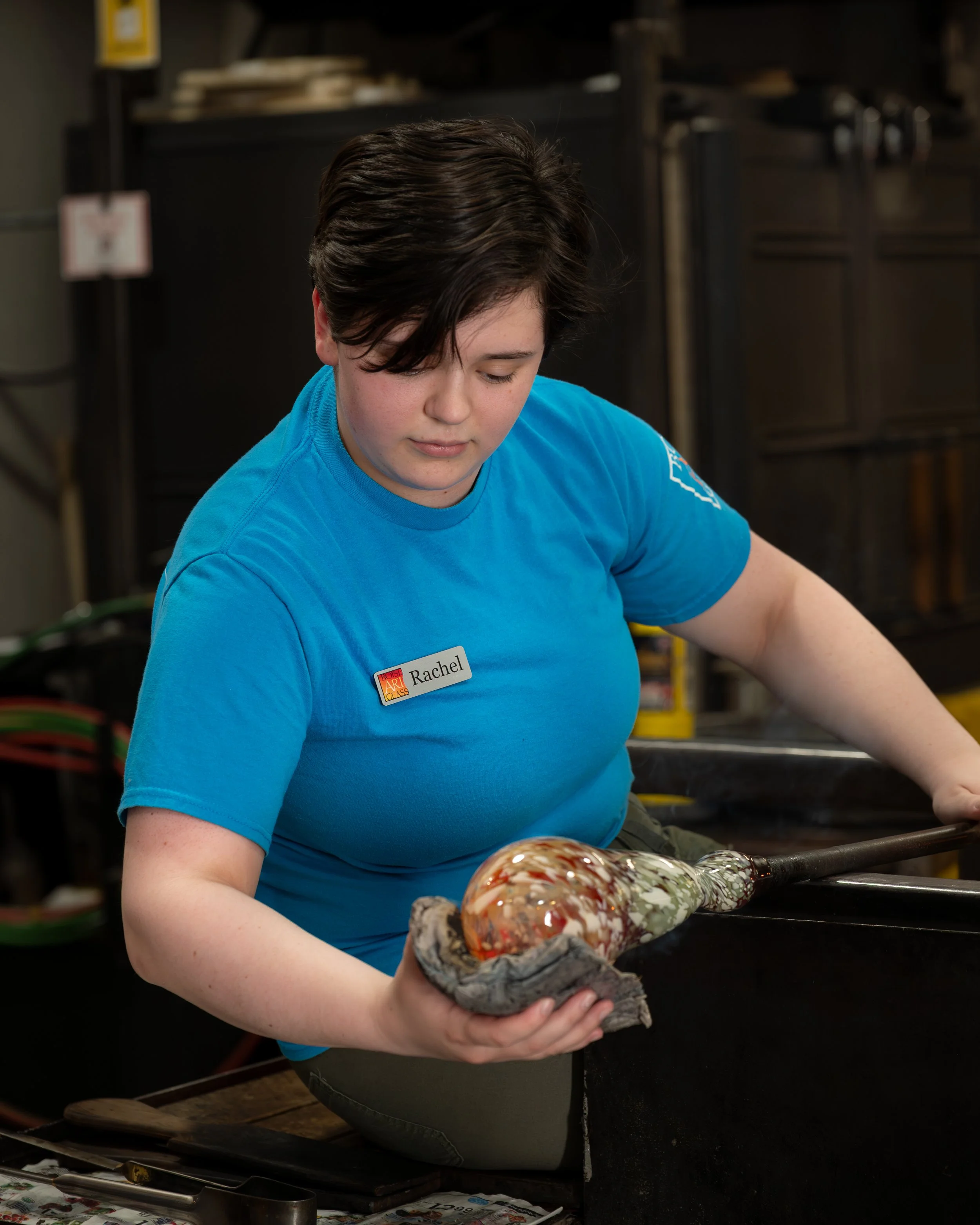 A glassblower wearing a blue shirt shaping a glass vase with multiple colors inside in a workshop setting.