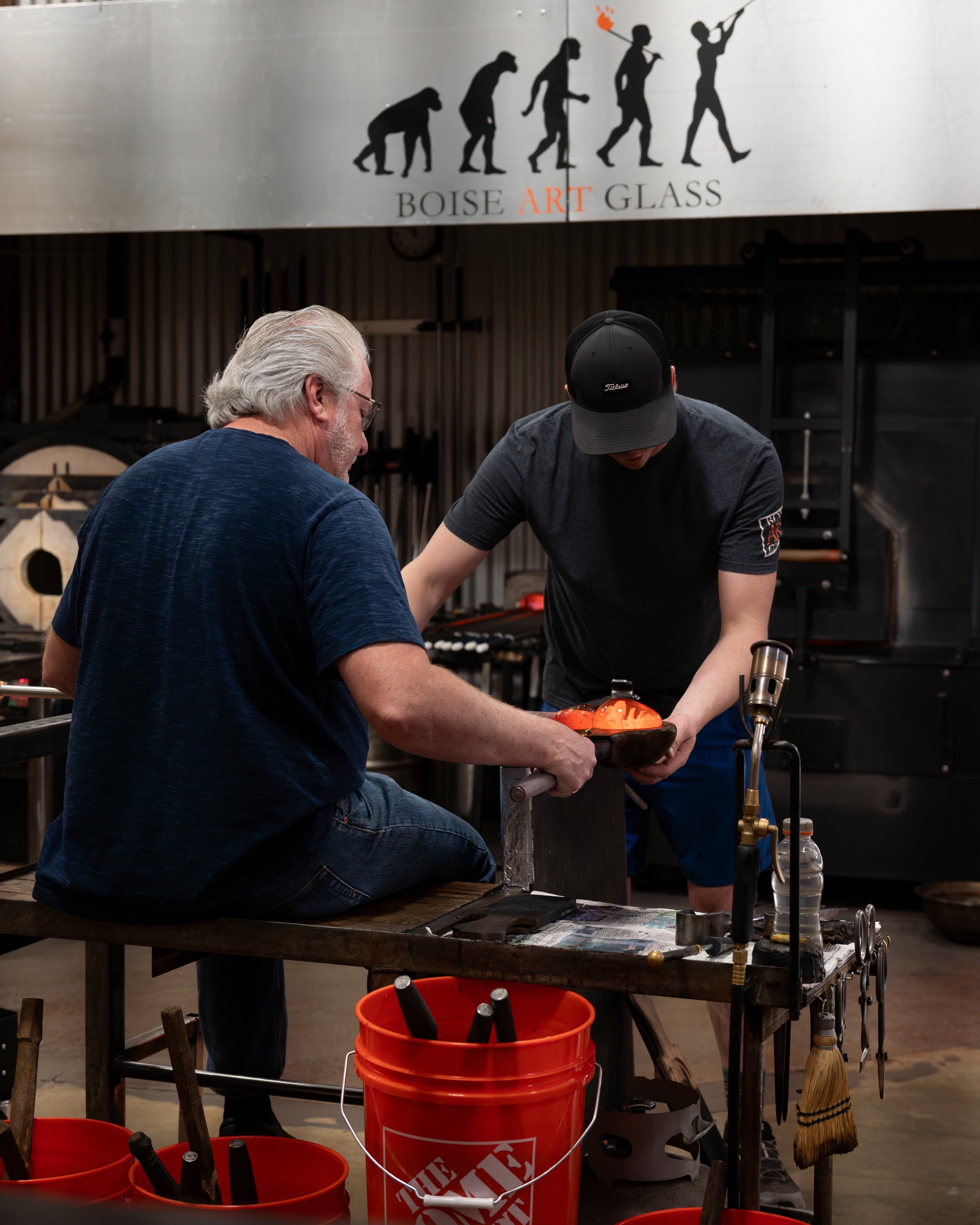 A glassblower teaching another man how to blowglass. There are orange buckets on the floor and a sign above with a progression of human evolution silhouettes and the words "Boise Art Glass."