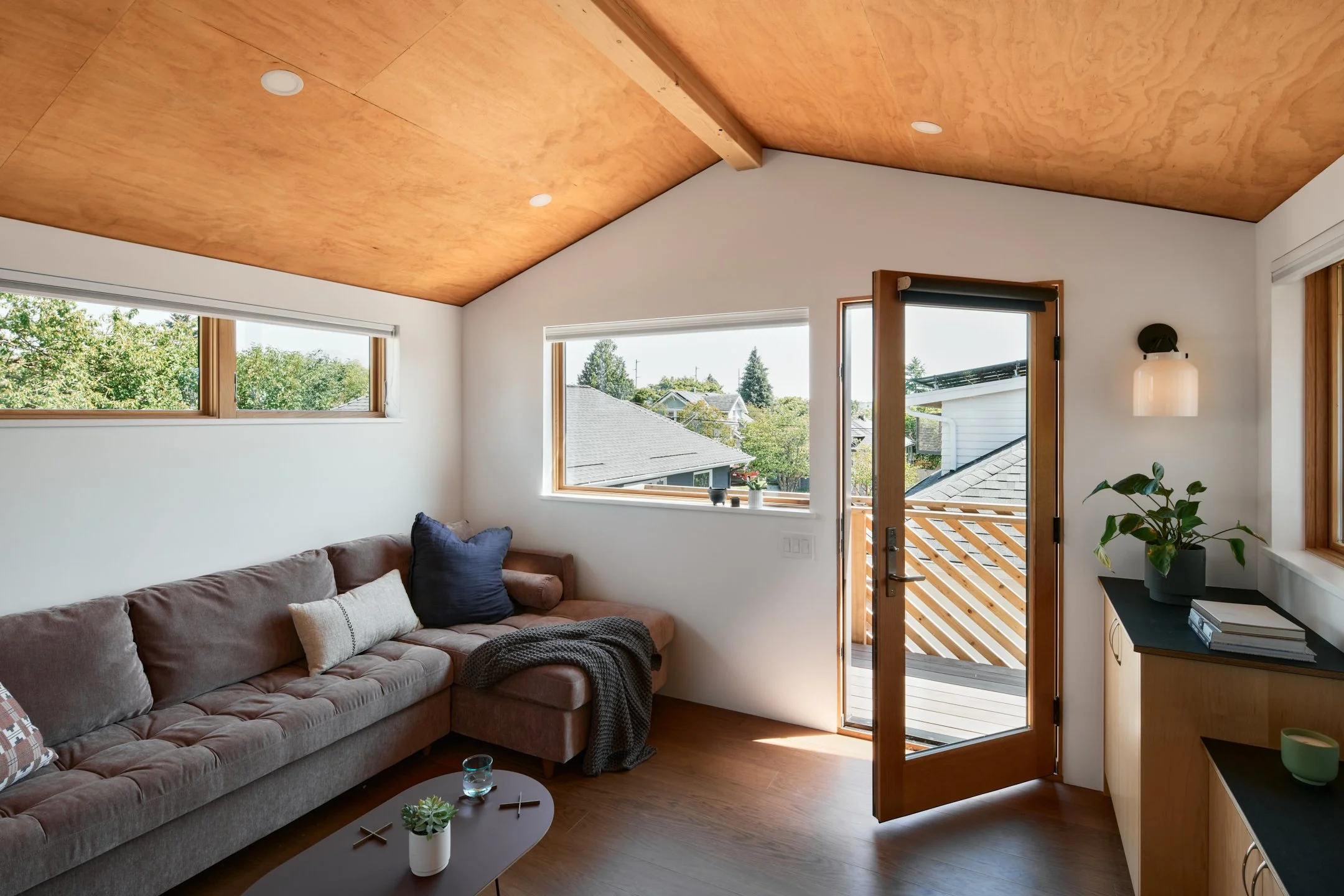 Living room with beige sectional sofa, throw pillows, small coffee table, potted plant, wall-mounted light, and large windows showing trees and neighboring houses.