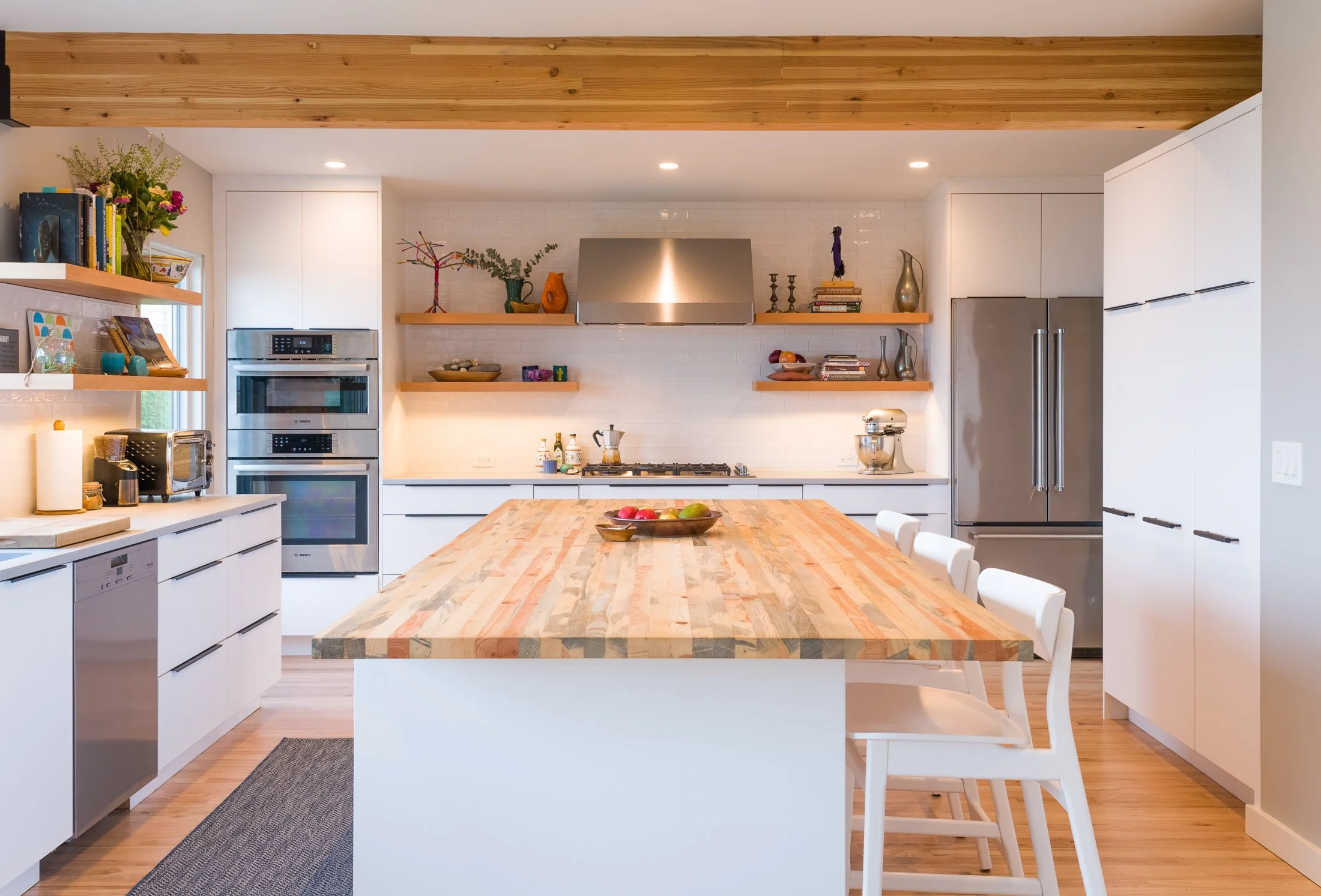 Modern kitchen with white cabinets, wooden open shelves, stainless steel appliances, a large wooden island with white chairs, and decorative items on shelves and countertops.