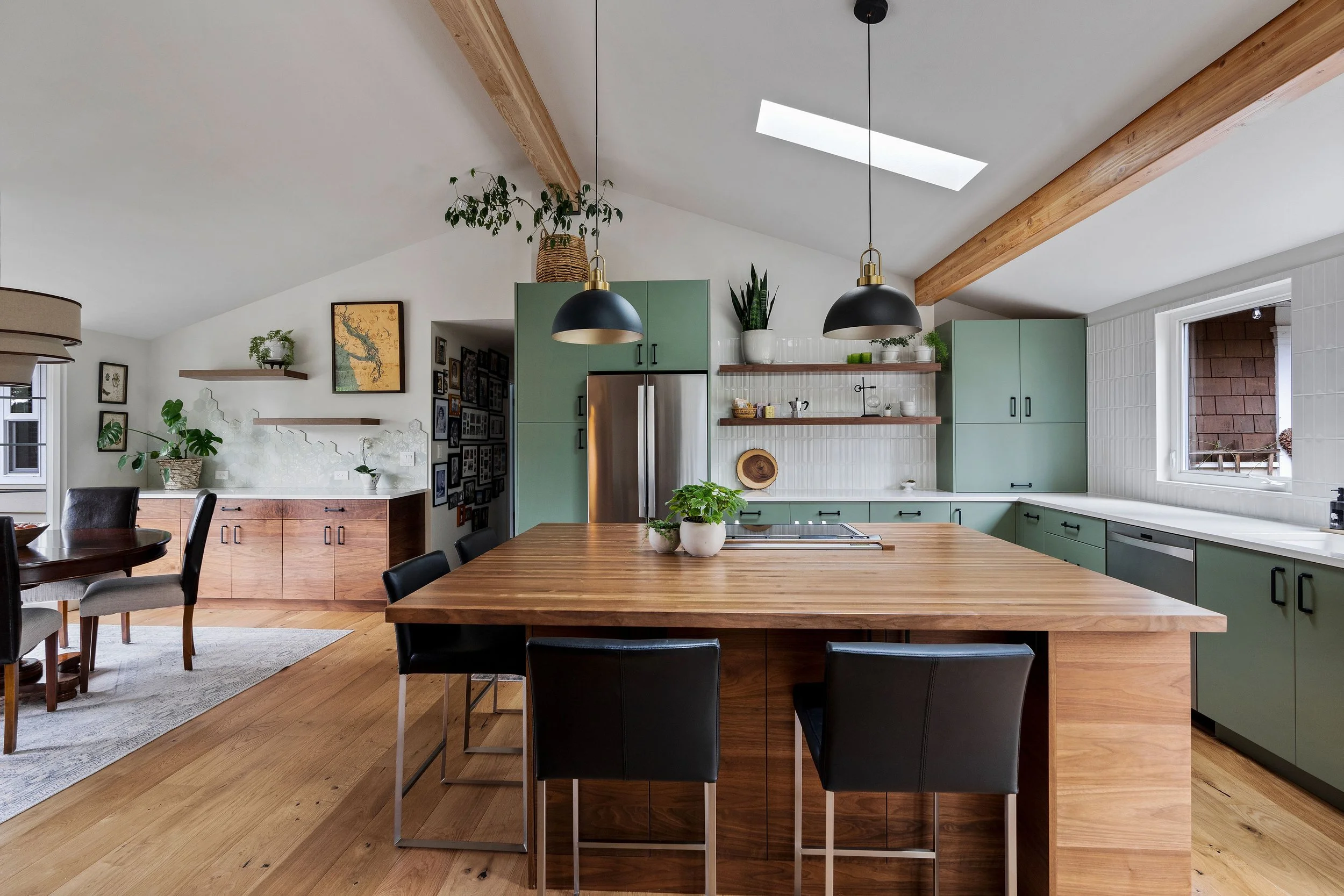 Modern kitchen with a large wooden island, black pendant lights, green cabinets, open shelving, and a skylight.