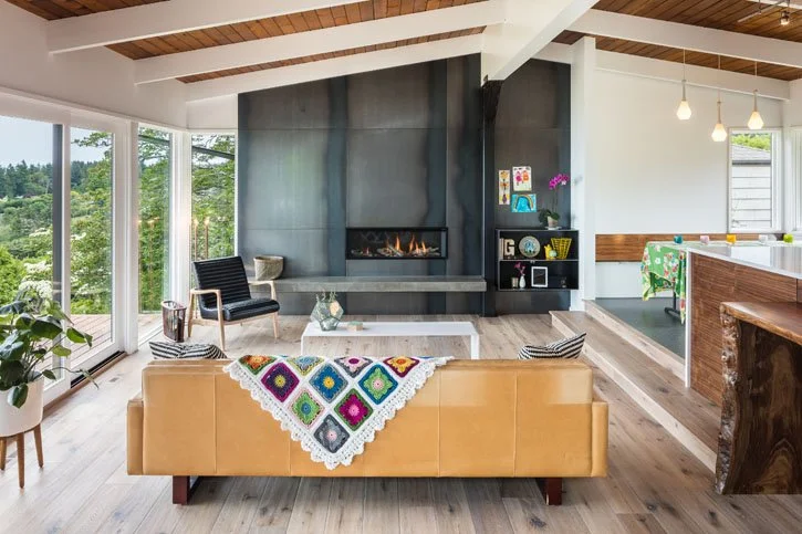 Living room with large windows, a black modern fireplace, a leather sofa with a colorful crochet blanket, a white coffee table, and wooden accents.