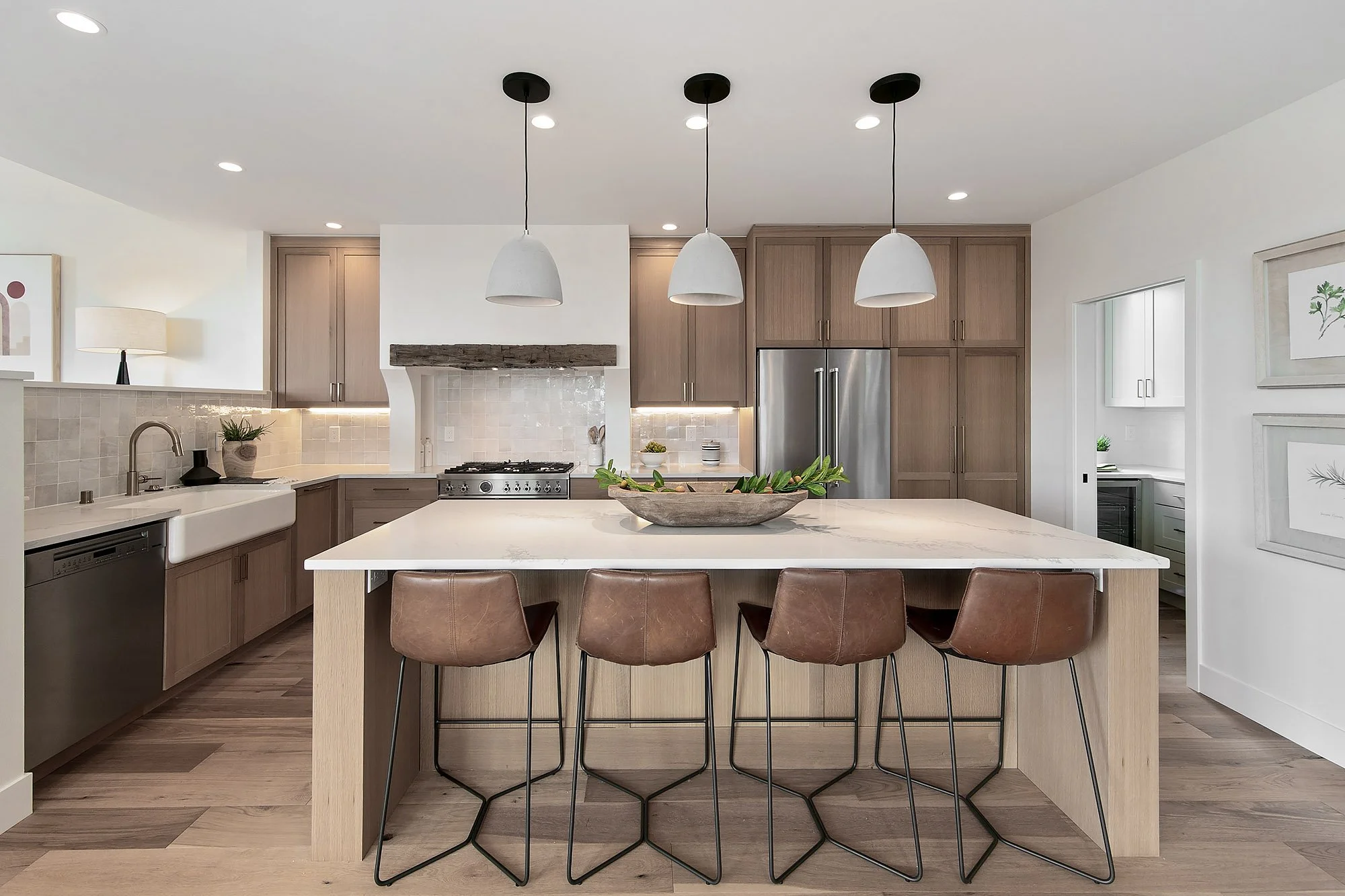 Modern kitchen with a large island, four brown leather chairs, pendant lights, wooden cabinets, stainless steel refrigerator, and a white backsplash.