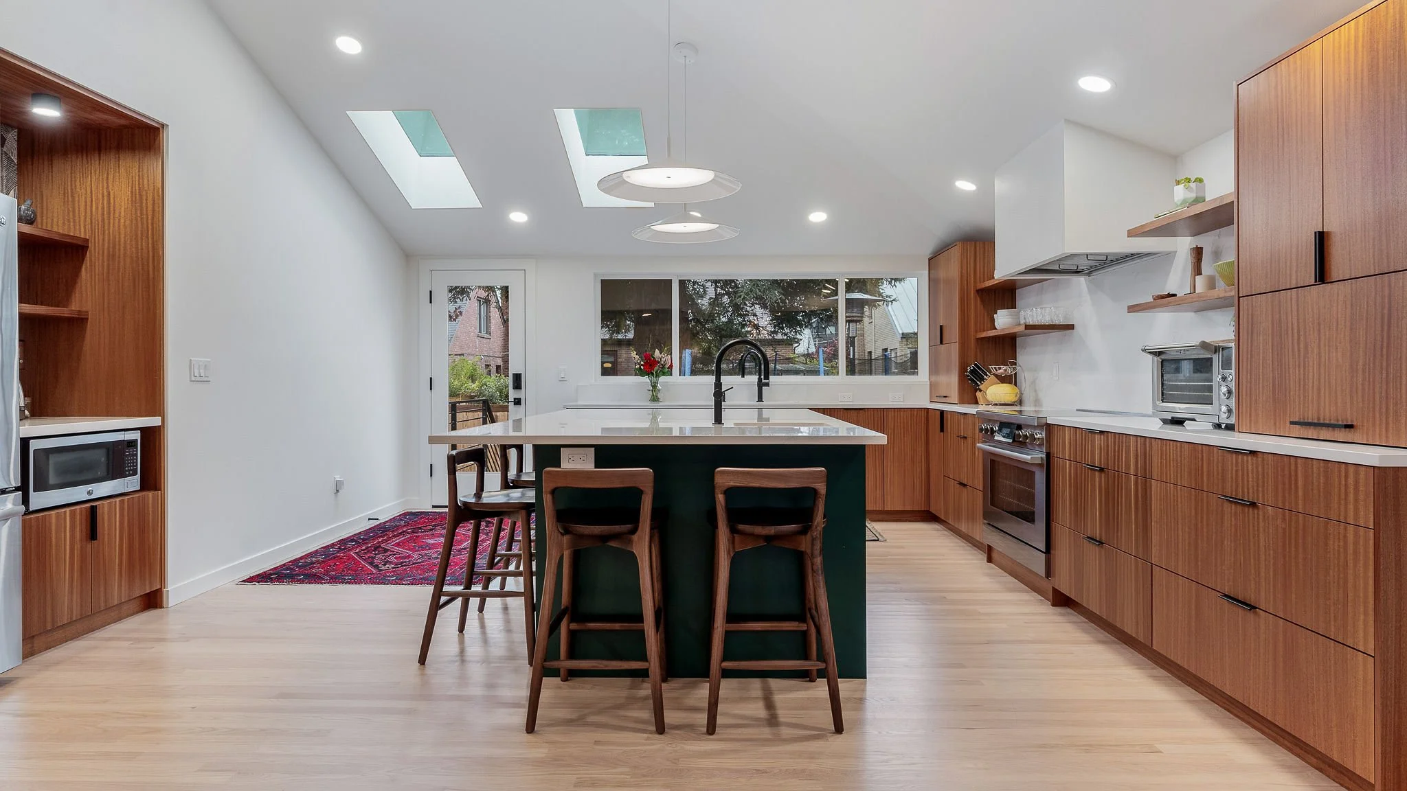 Modern kitchen with white walls, wooden cabinets, a large island with a green base, three wooden chairs, skylights, and a window showing trees outside.