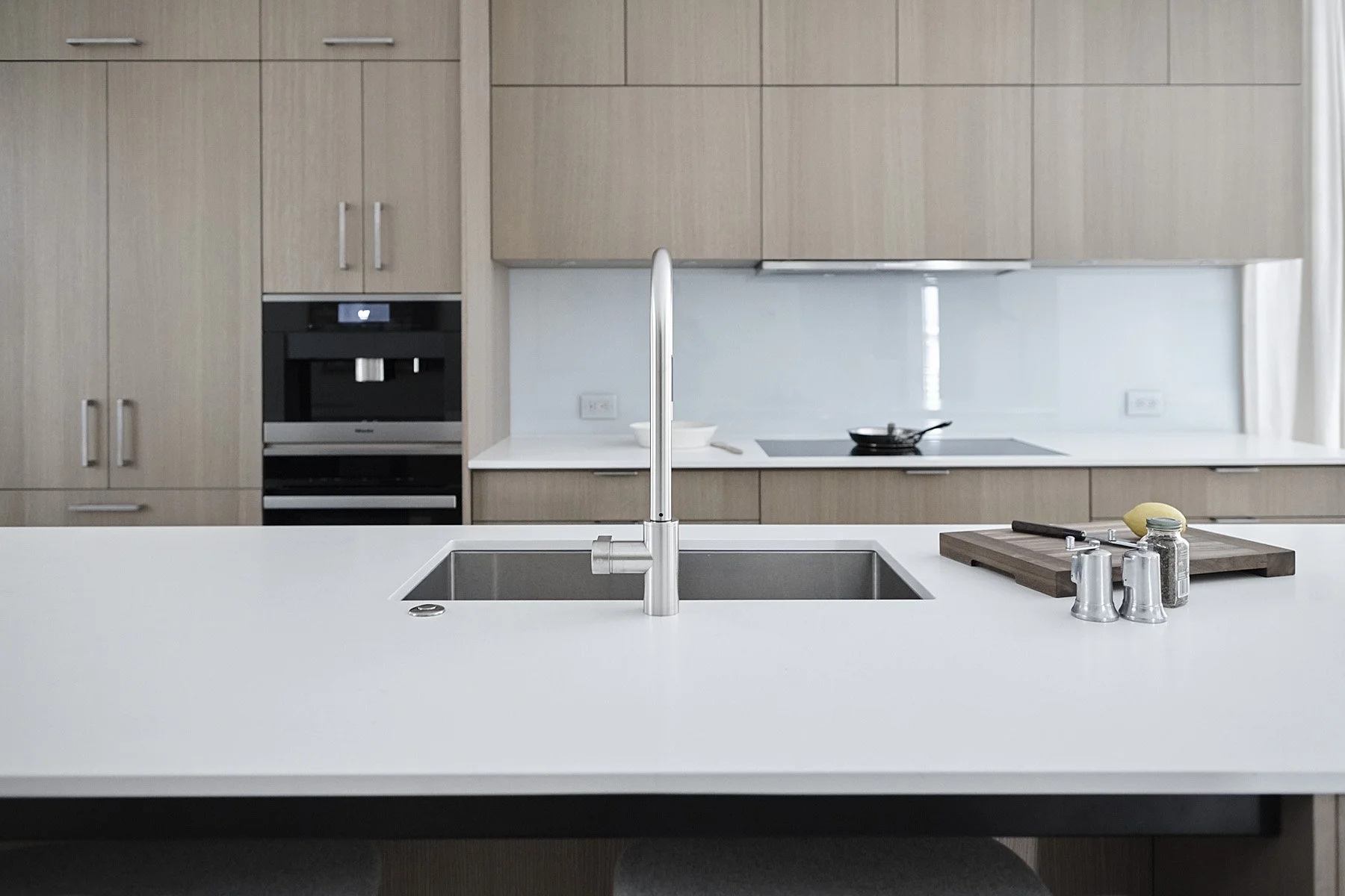 Modern kitchen with light wood cabinets, white countertop, and a stainless steel sink with a tall faucet. Kitchenware including a cutting board, knife, and spice jars are on the countertop. A built-in microwave and oven are integrated into the cabine