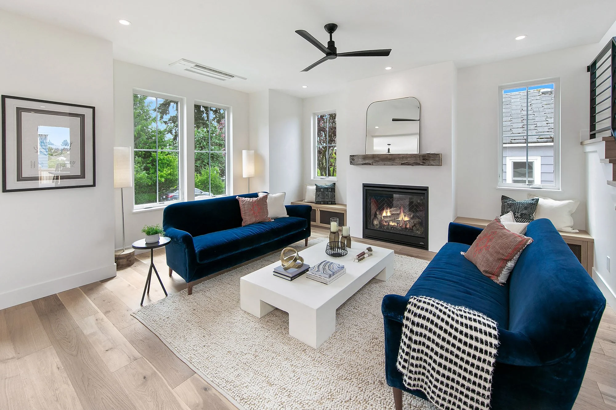 Modern living room with two blue velvet sofas, a white coffee table, a fireplace with a wooden mantel, a large mirror above the fireplace, three tall windows, and minimal decor.