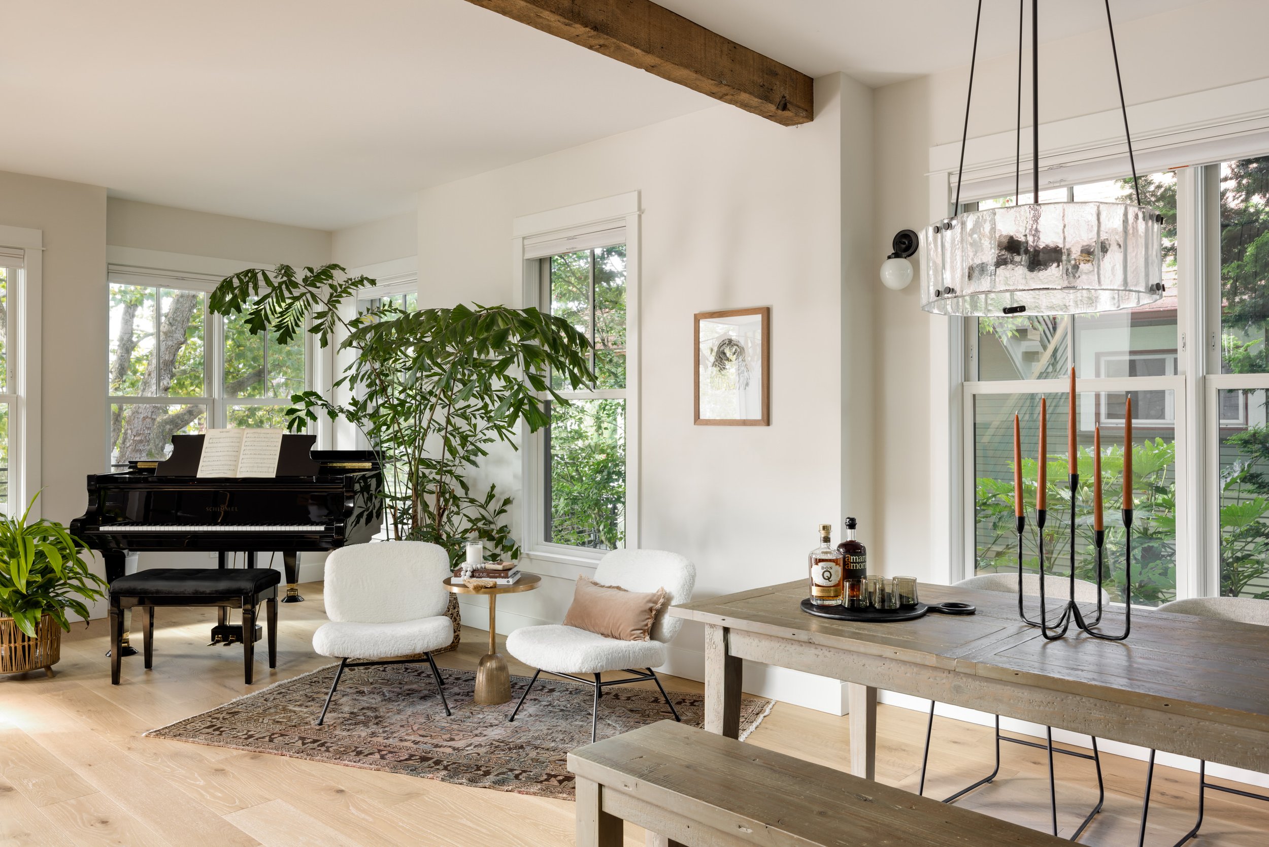 Living room with white walls and hardwood floors, featuring a black grand piano, an indoor plant, two white chairs, and a wooden table with bottles and glasses. Large windows reveal greenery outside, and a modern chandelier hangs above a dining table.