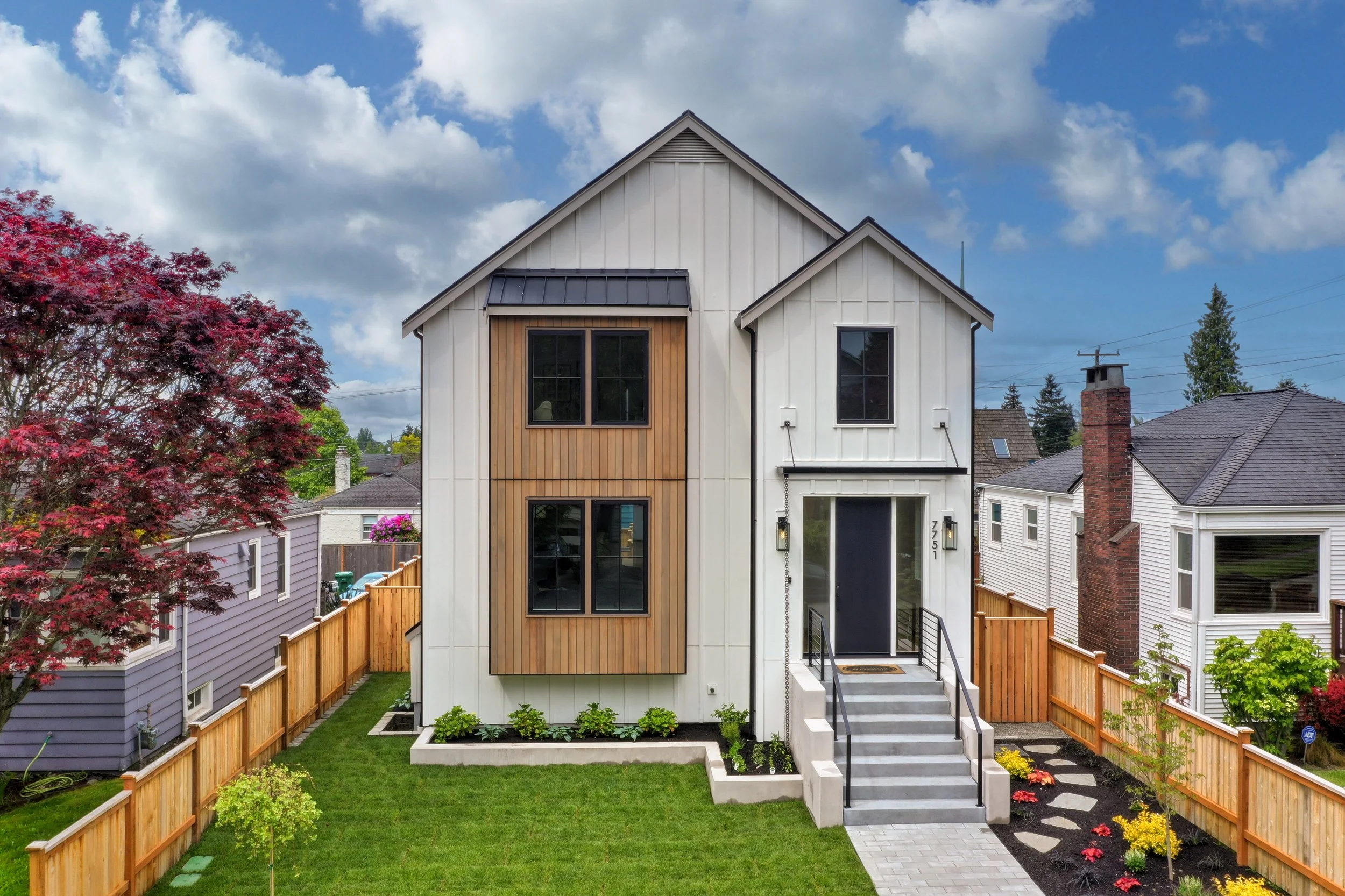 Modern two-story house with white siding, wood accents, black window frames, gray steps, a small front yard with green grass and flower beds, wooden fencing, and neighboring houses. A tree with red leaves is on the left, under a partly cloudy sky.