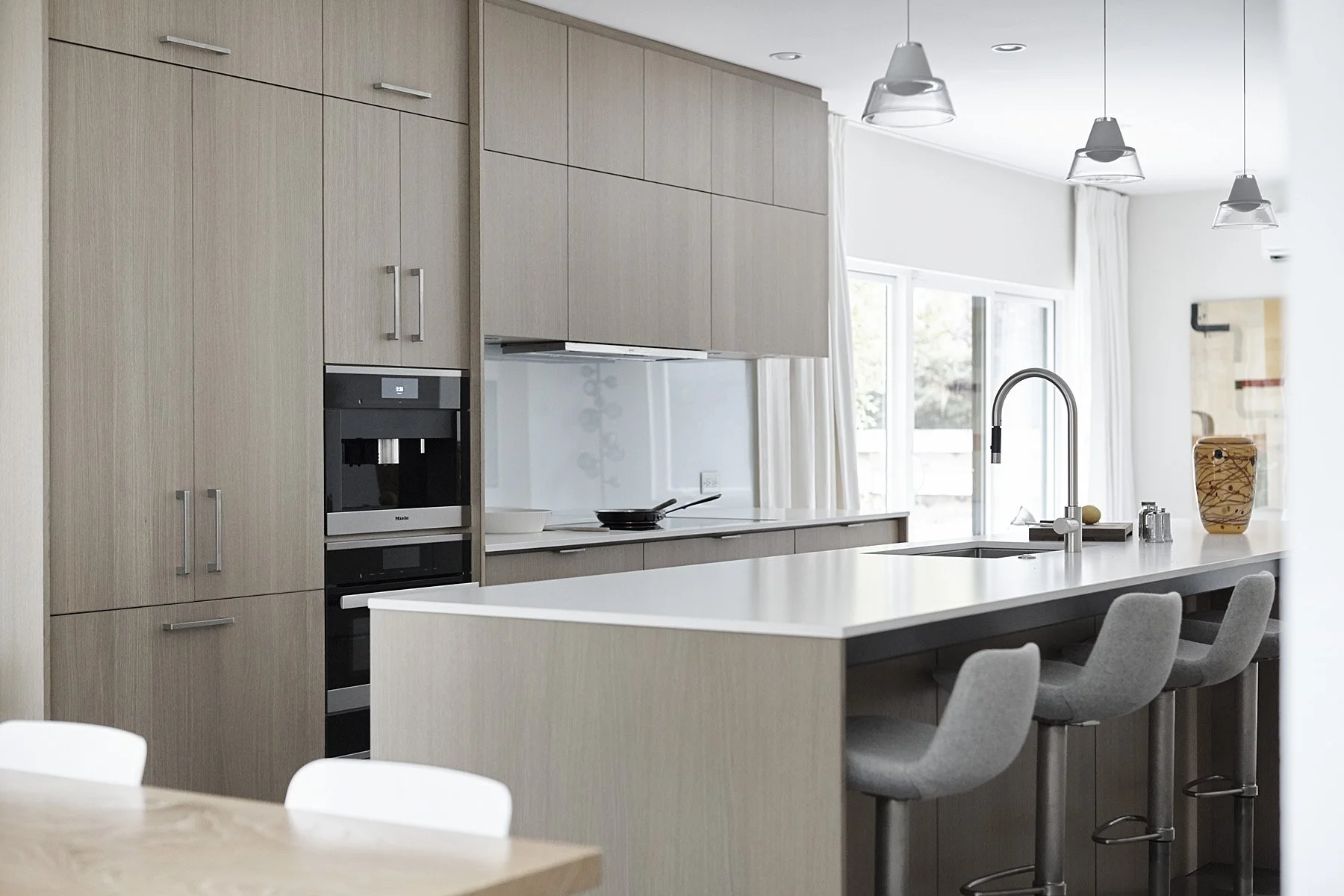 Modern kitchen with beige cabinets, white island, gray bar stools, and large windows with white curtains.
