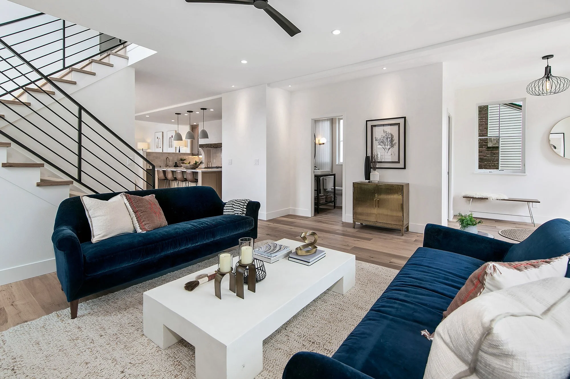 Modern living room with navy velvet sofas, a white coffee table, and decorative candles and books, with a staircase and kitchen area in the background.