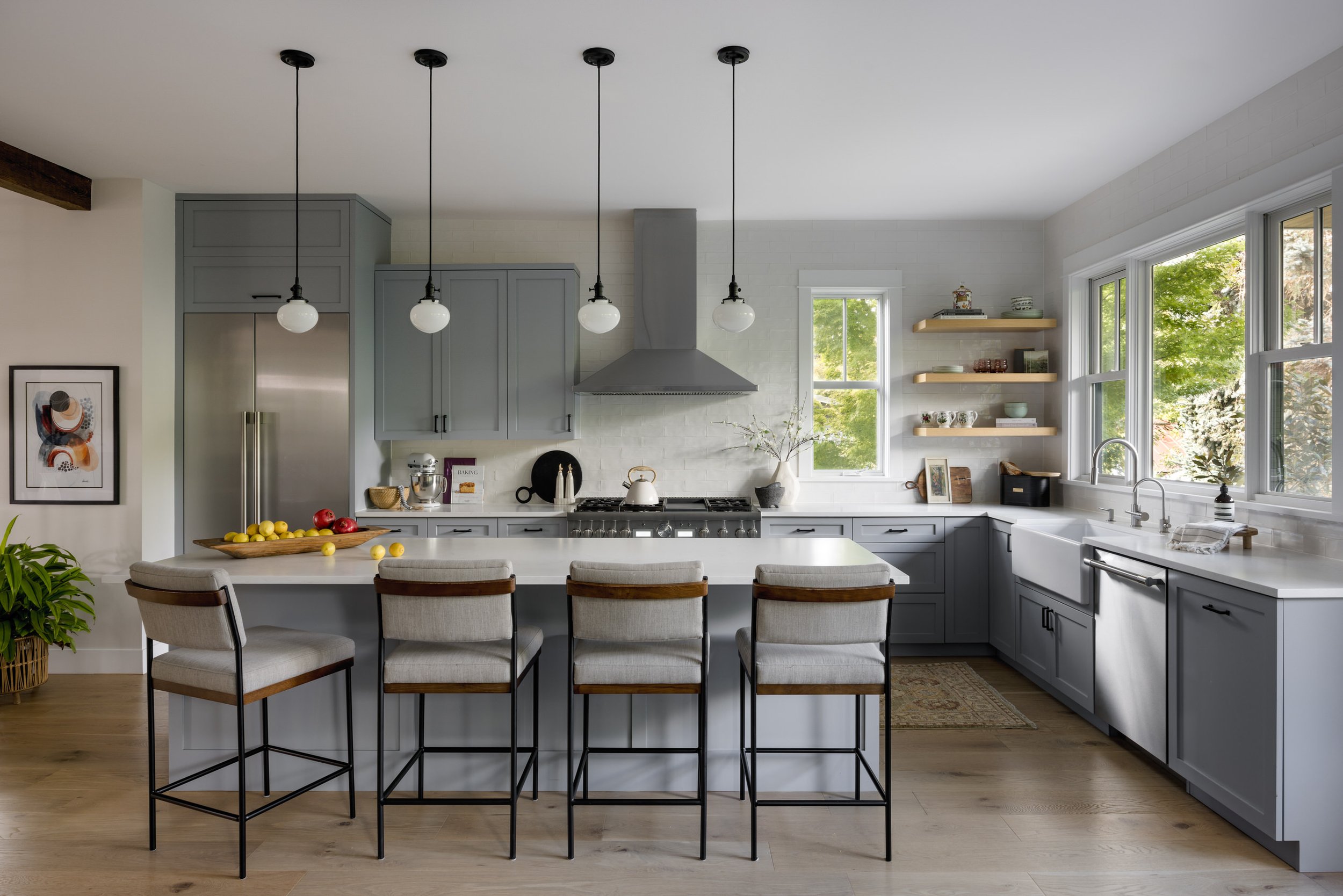Modern kitchen with gray cabinets, white countertops, a kitchen island with four bar stools, large windows, and a stainless steel range hood.