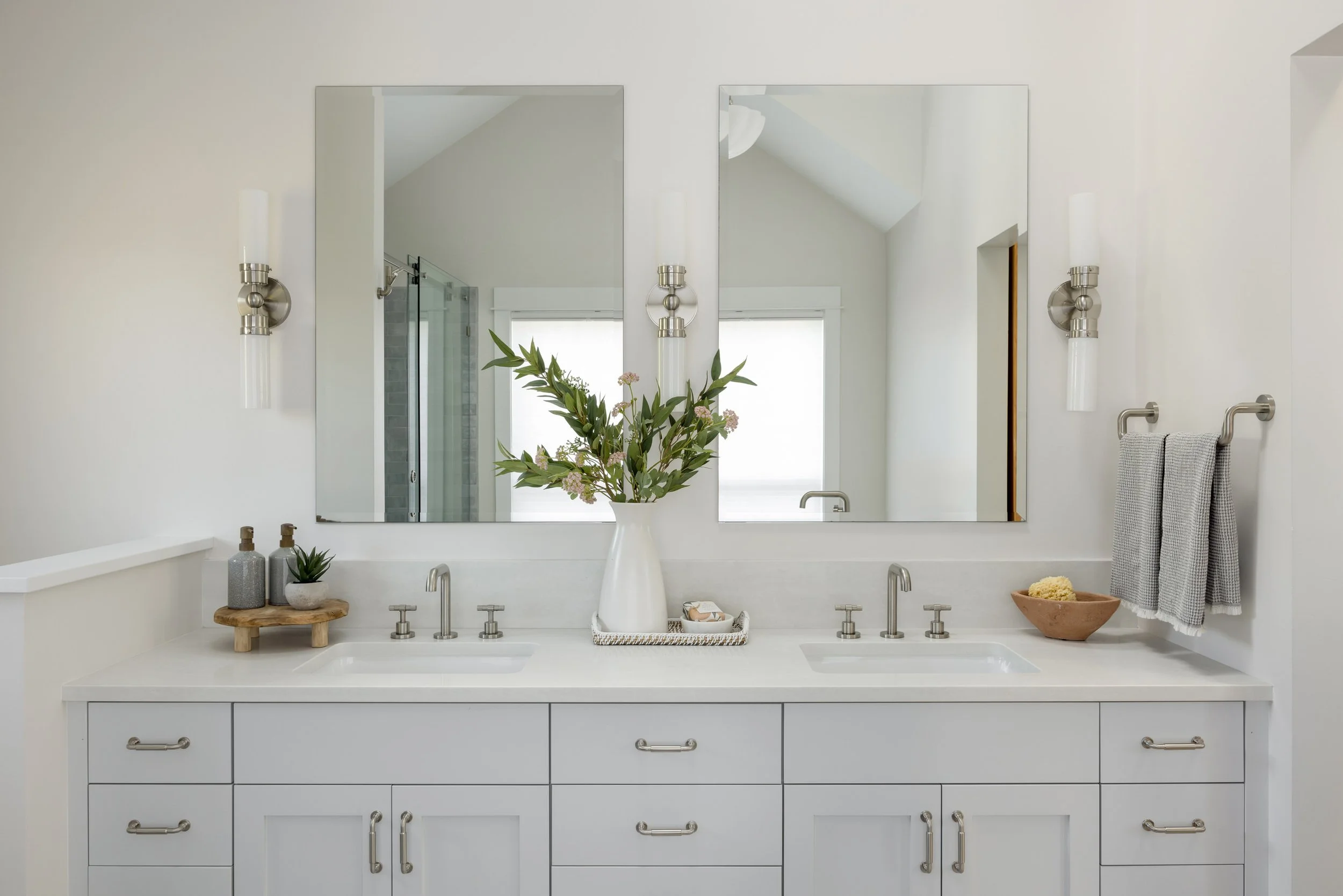 A double-sink bathroom vanity with two large mirrors, topped with a white vase containing greenery and purple flowers, and decorated with small potted plants, soap dispensers, and a bowl of sponge.