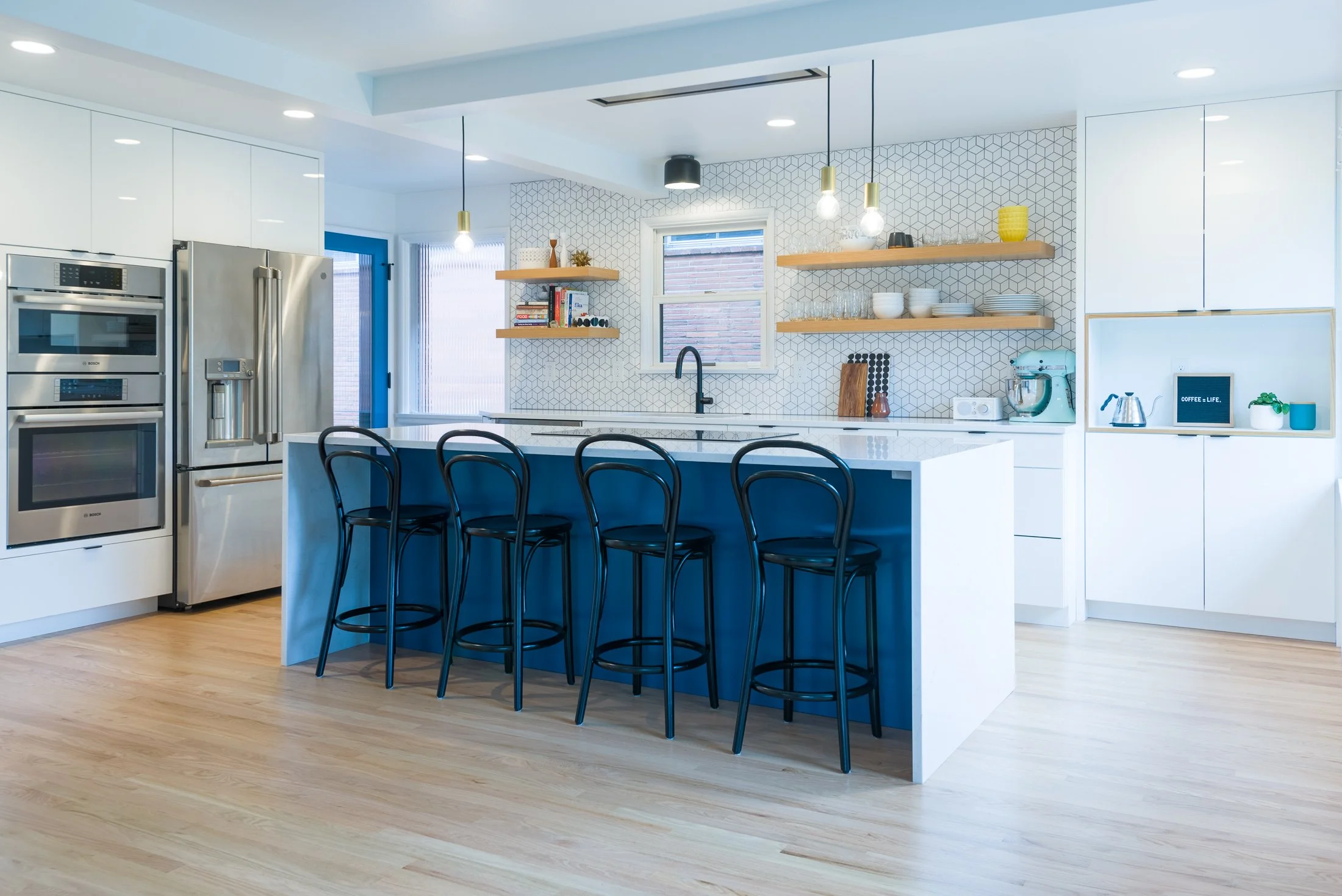Modern kitchen with white cabinets, a large kitchen island with a blue base, four black chairs, stainless steel appliances, open wooden shelves with dishes and books, and pendant lights.