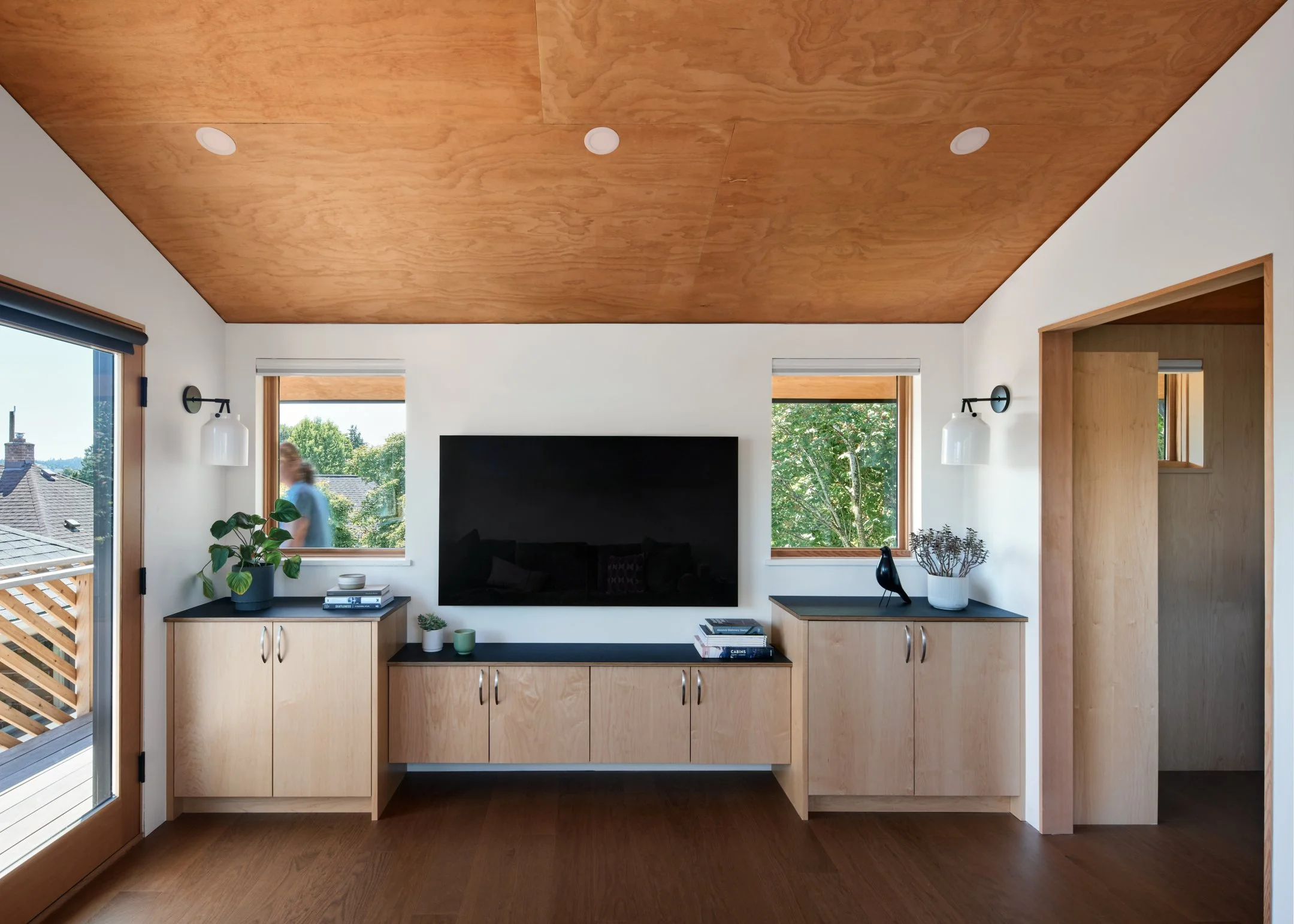 Living room interior with wooden ceiling, white walls, two windows, a wall-mounted TV, beige cabinets, indoor plants, decorative items, and a door leading to an outdoor balcony.