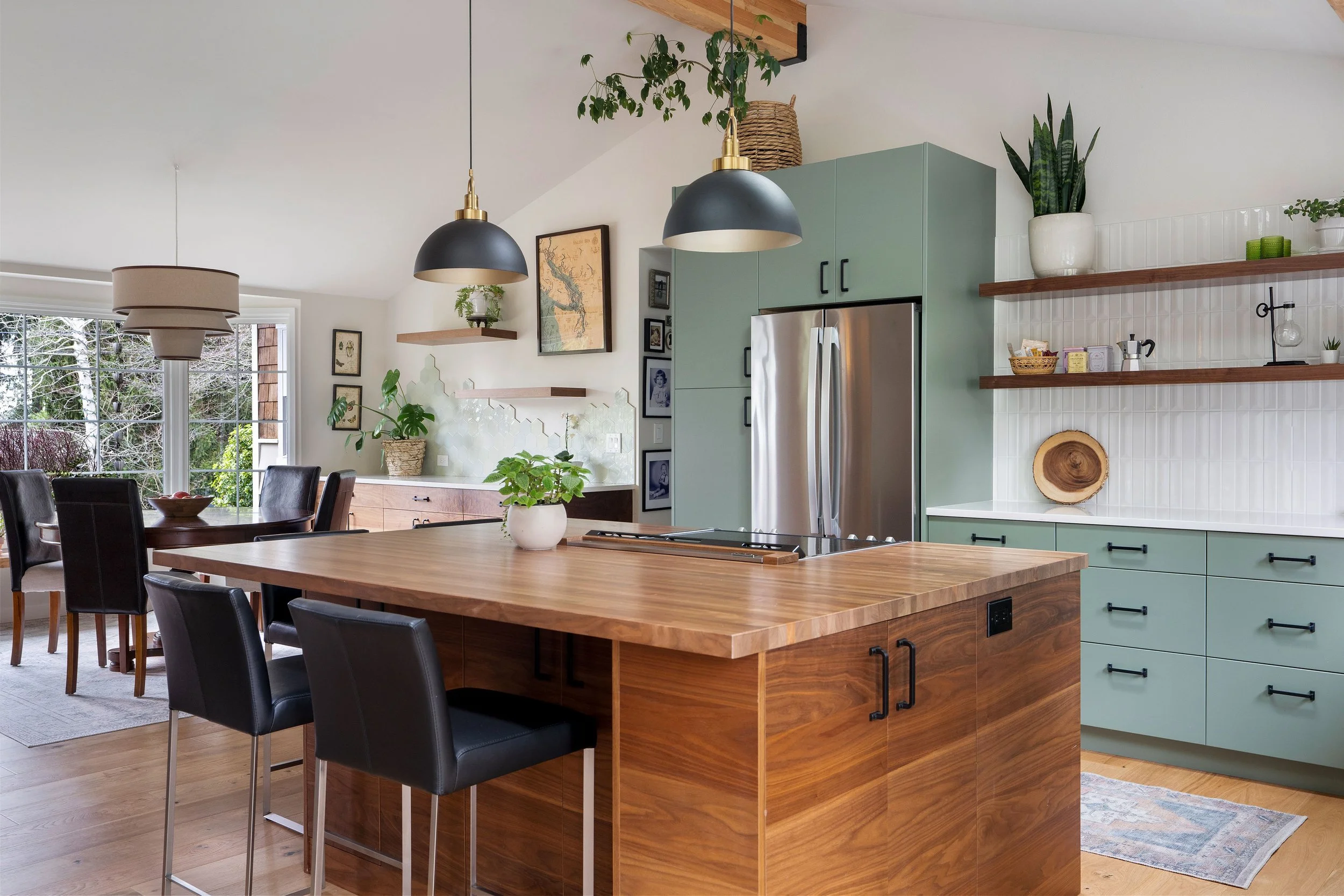Modern kitchen with a wooden island, green cabinetry, open shelves with plants and decorations, stainless steel refrigerator, black pendant lights, and a dining area near large windows showing a snowy outdoor scene.