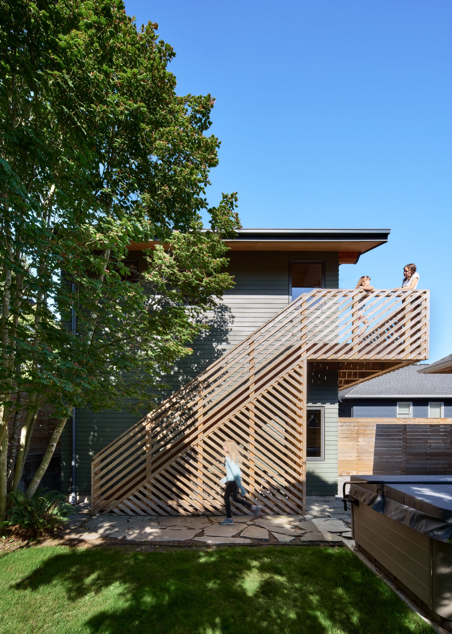 Three children are on a second-story balcony of a modern house; one child is walking on the patio below, while a girl talks to another girl on the balcony. The house features wood accents, and there is a large green tree on the left side under a clea